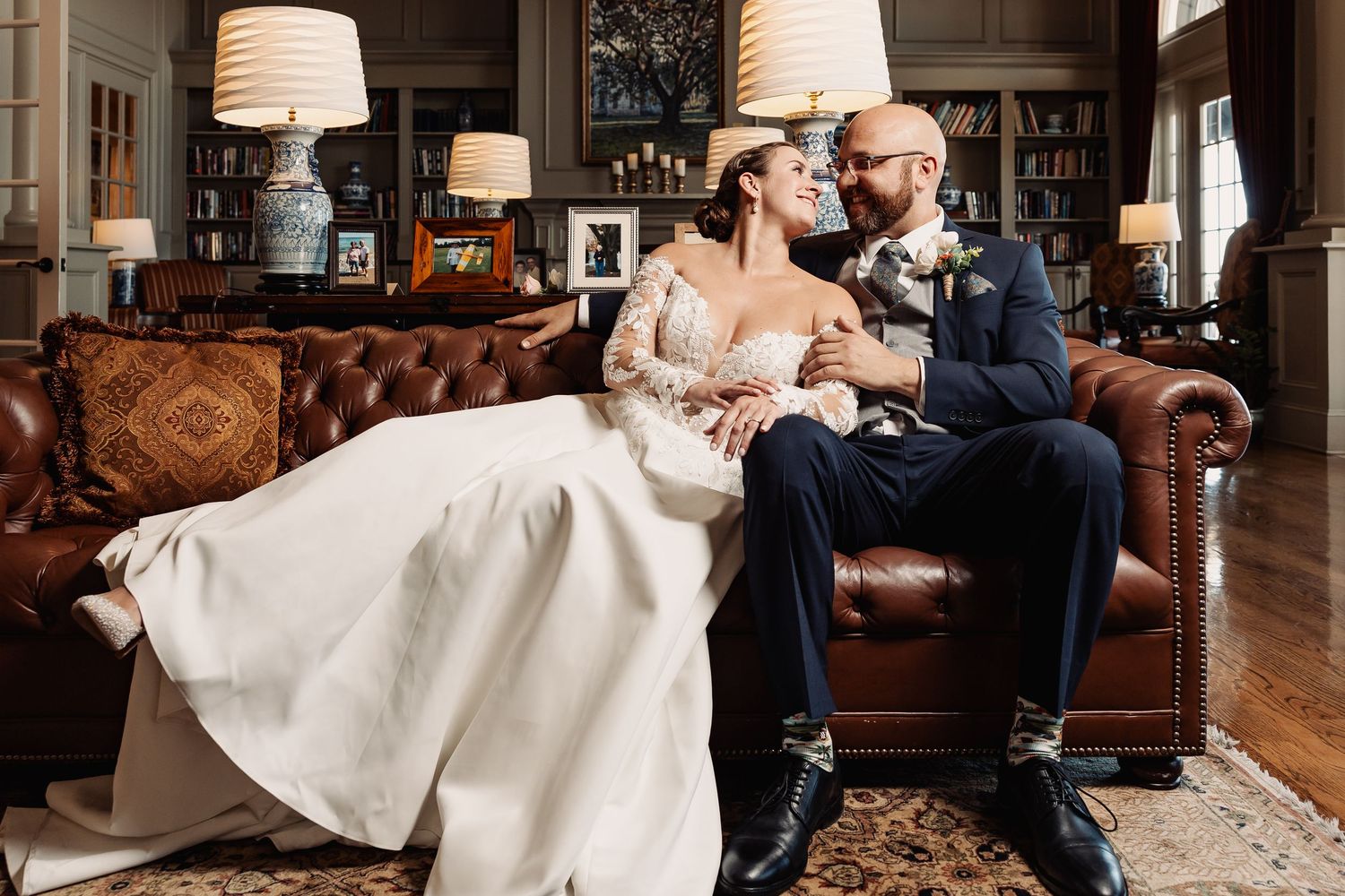 Bride and groom share romantic moment on leather sofa in elegant library on wedding day.