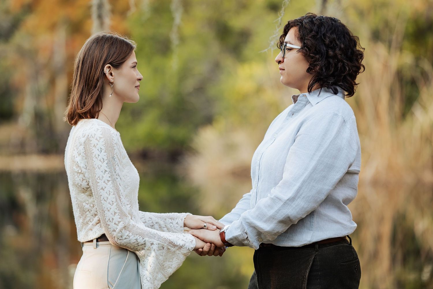 Two women holding hands facing each other near a reflective pond during an outdoor ceremony.