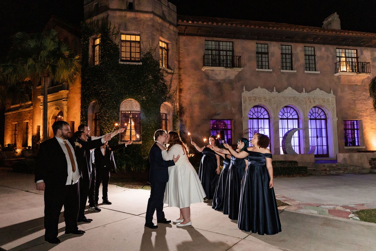 Wedding party holds sparklers during a nighttime send-off outside a beautifully lit historic mansion.
