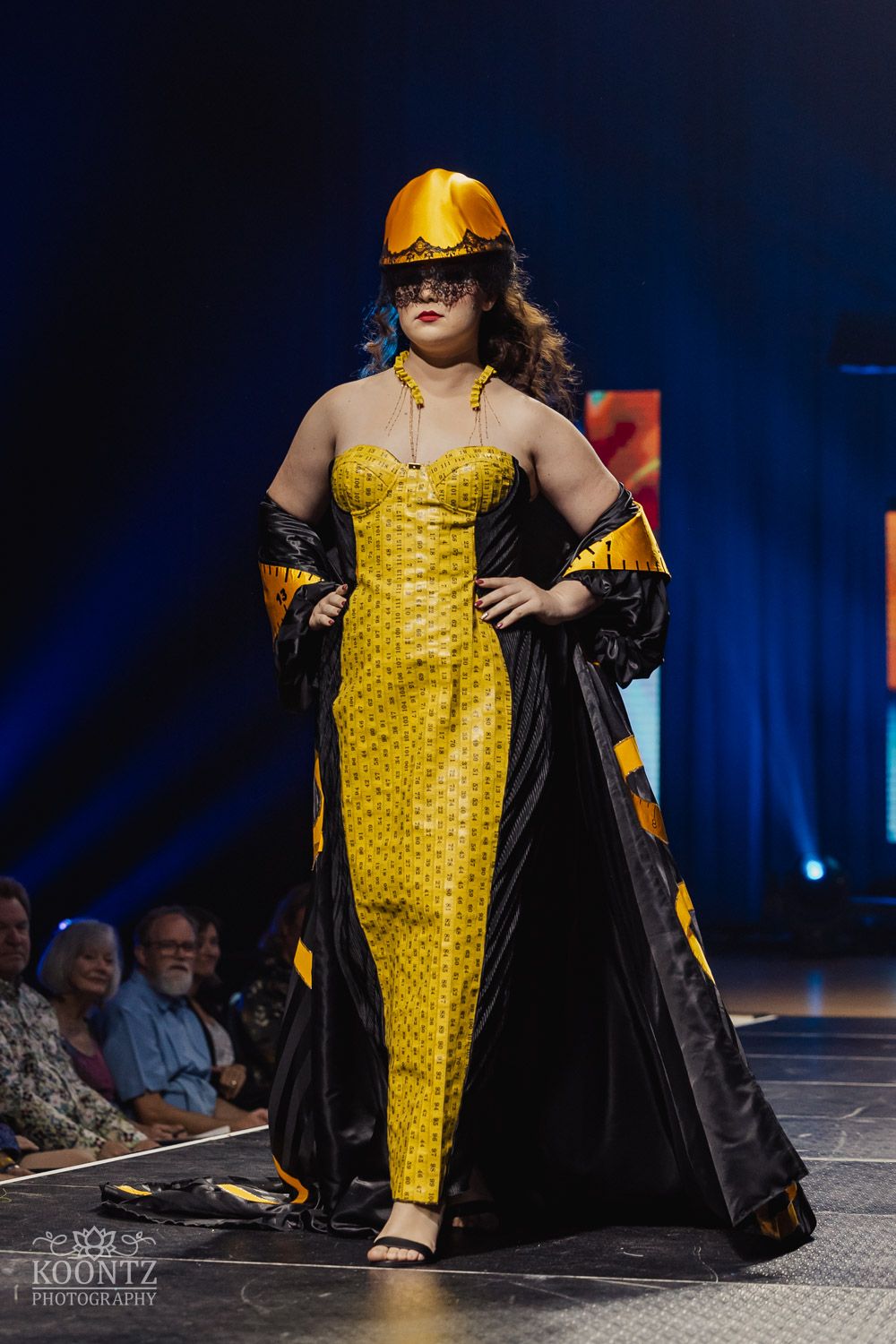 Model walks runway in striking yellow quilted jumpsuit with dramatic black cape at fashion show.