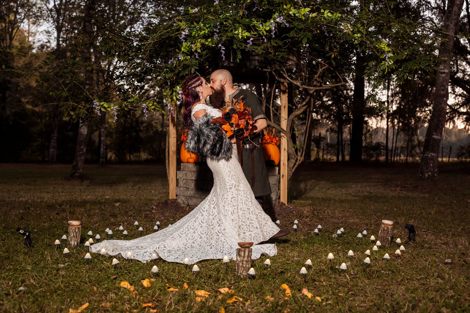 Romantic wedding dance under trees with glowing lights and white flower petals scattered on grass.