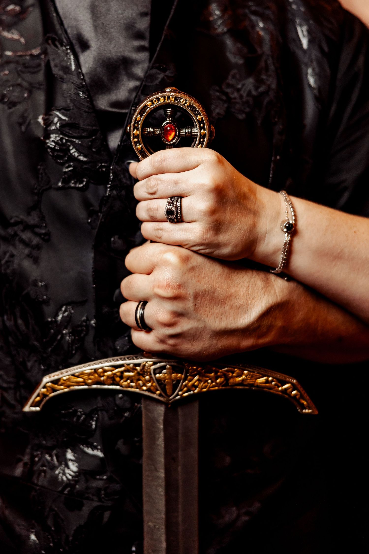 Close-up of hands gripping ornate gold and red gemstone sword hilt with rings and bracelet.