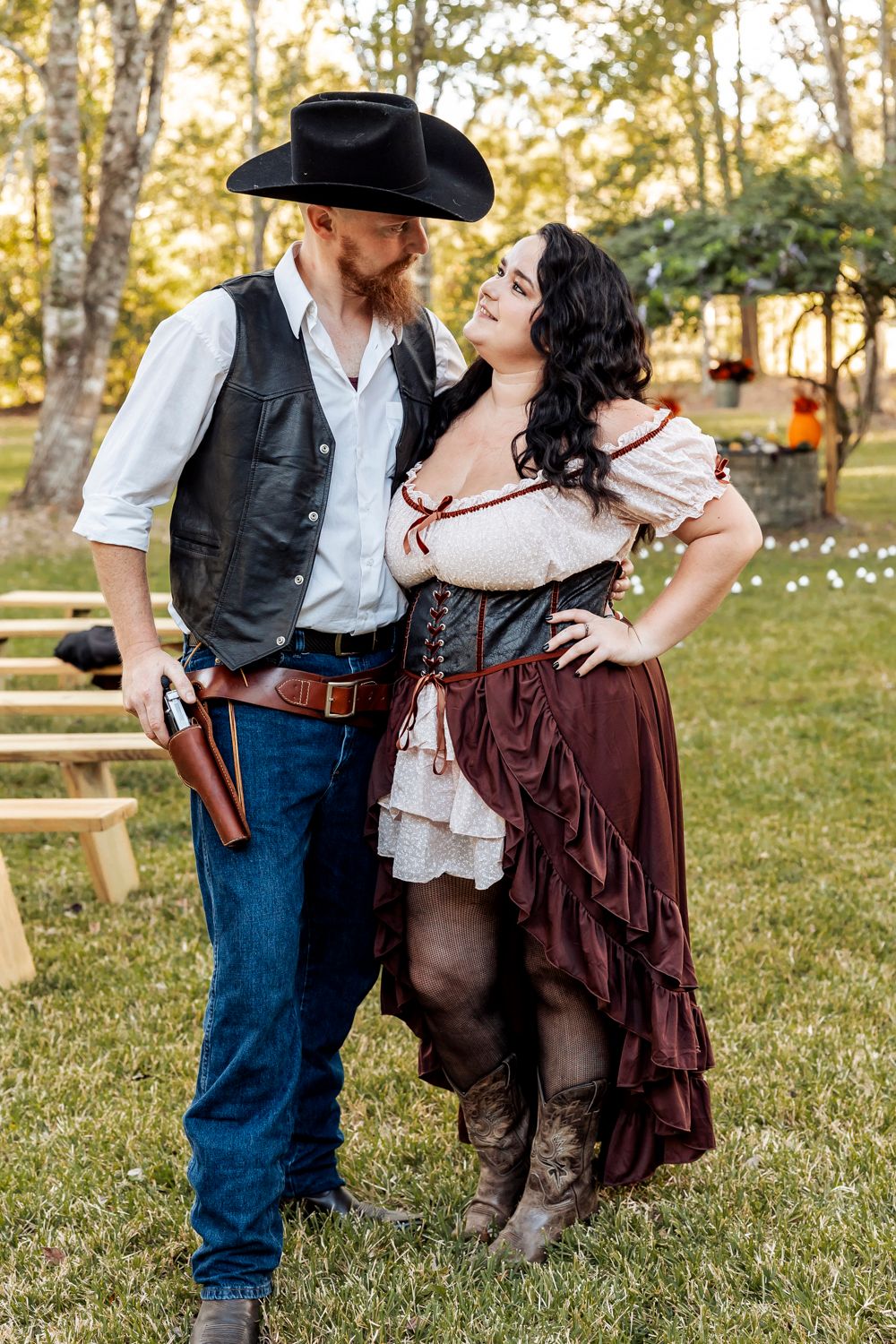 Couple in Western costumes pose outdoors, man in cowboy hat and vest, woman in corset saloon dress.