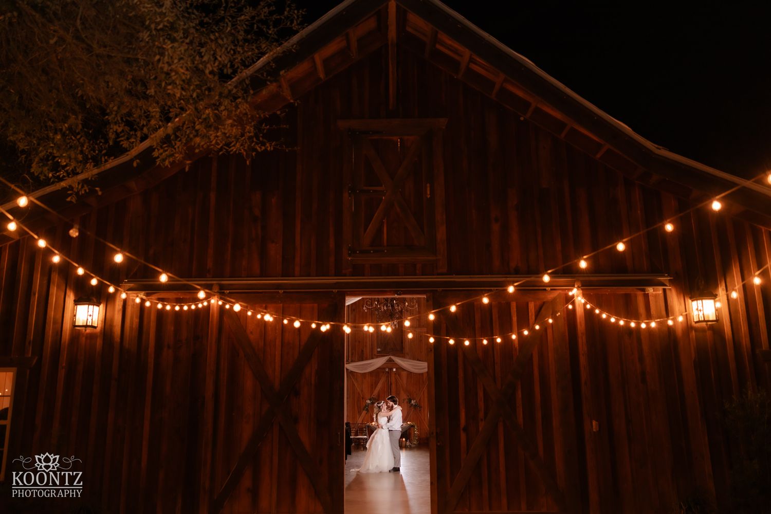 Newlyweds silhouetted in doorway of rustic red barn decorated with string lights at night wedding.