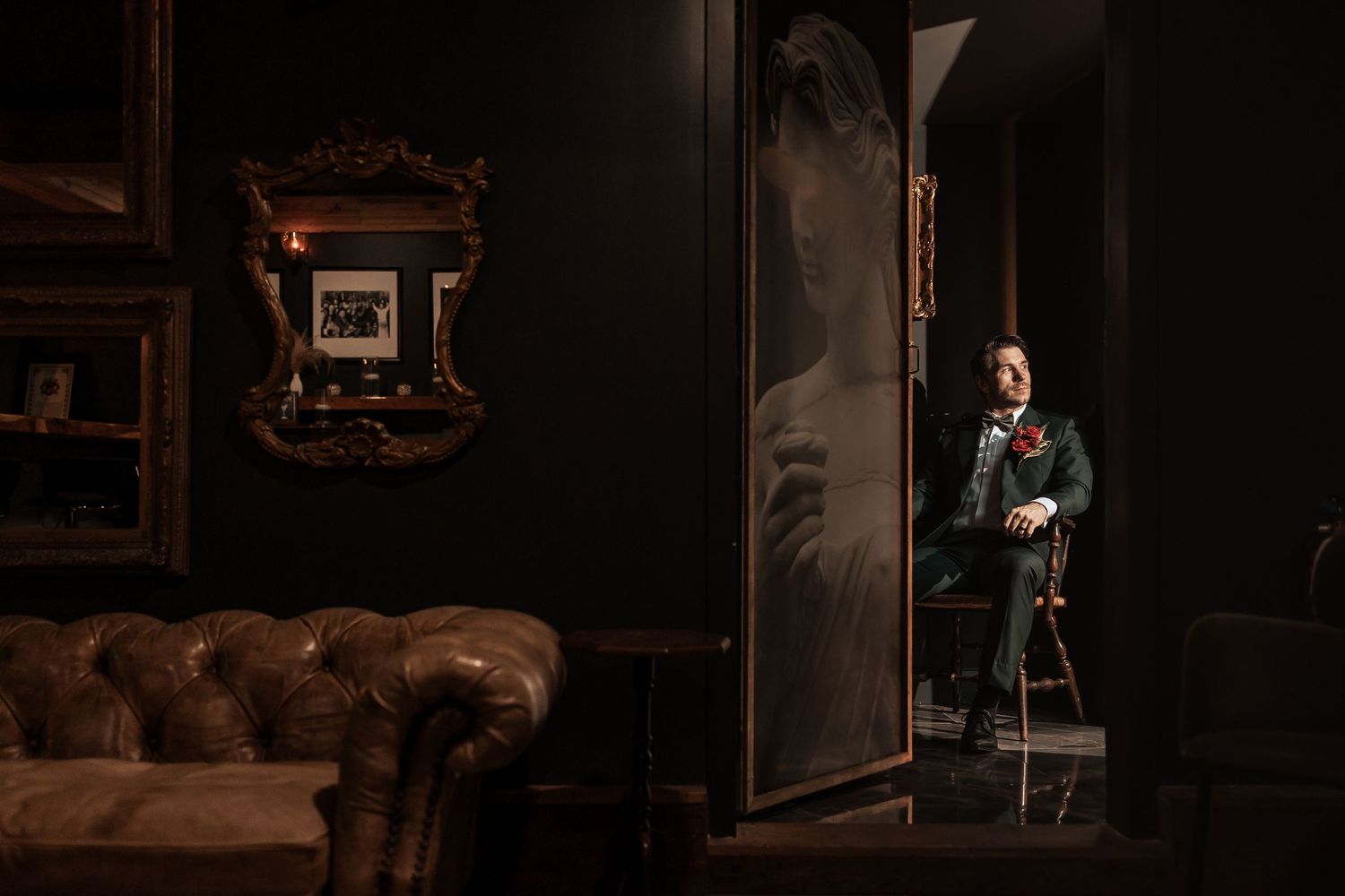 Groom in dark suit standing in a dimly lit elegant room with vintage decor and leather furniture.