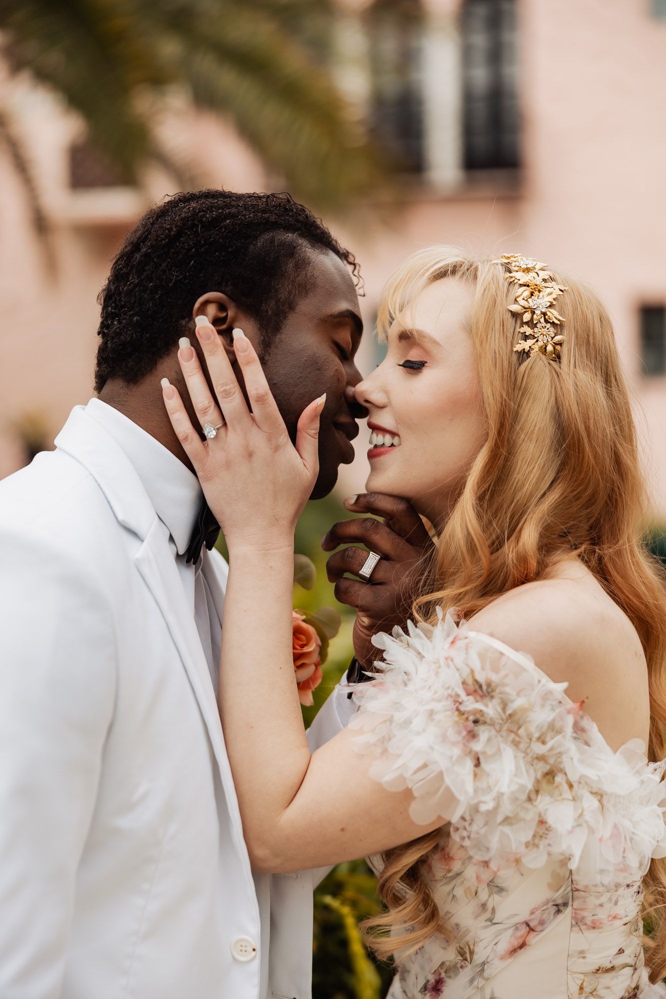 Romantic interracial bride and groom sharing intimate almost-kiss moment outside pink wedding venue.
