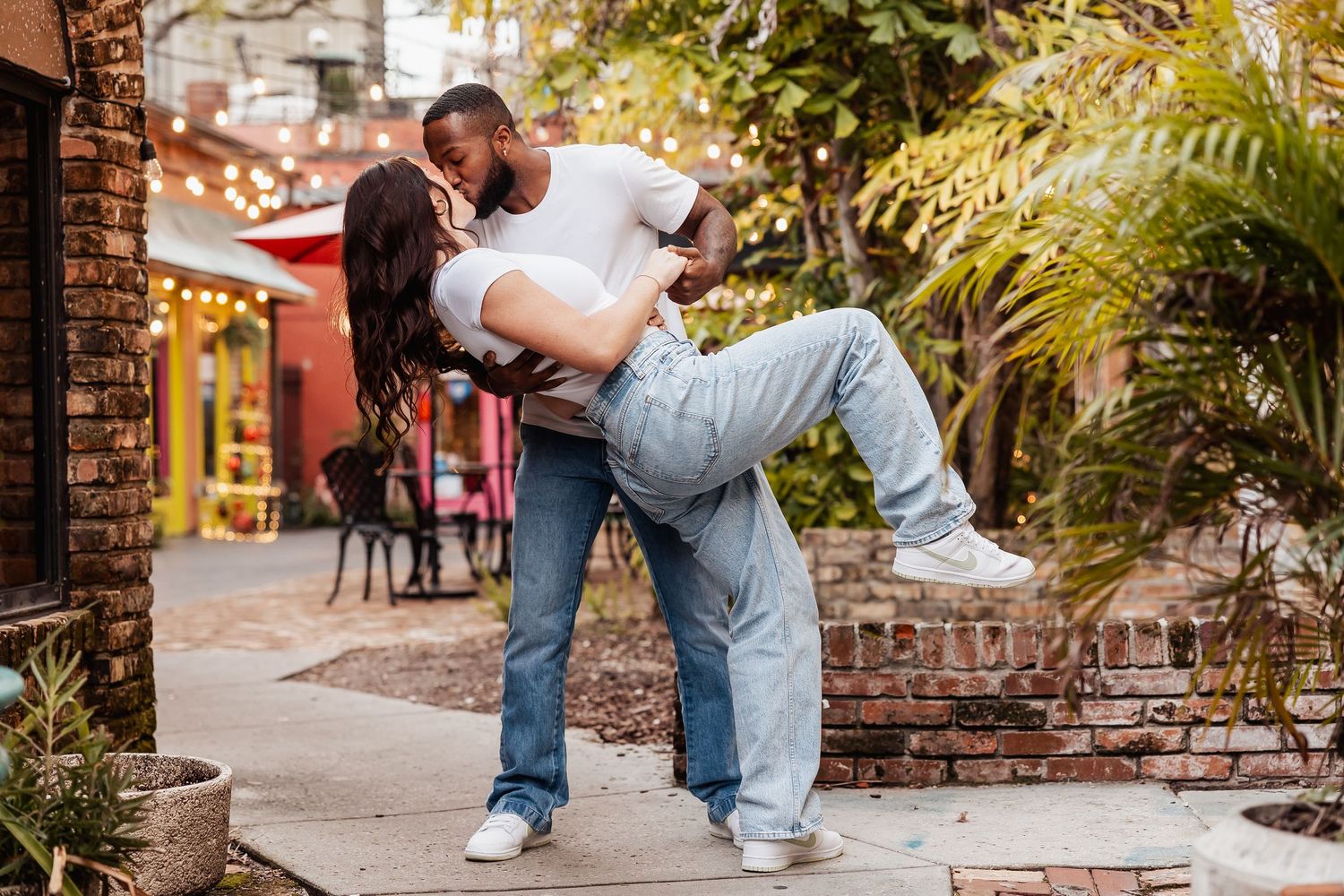 Couple sharing a romantic dip kiss outside a lush brick courtyard with string lights.