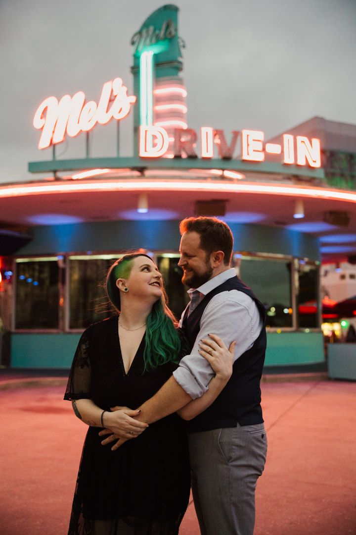 Smiling couple embracing in front of the glowing neon Mel's Drive-In sign at dusk.