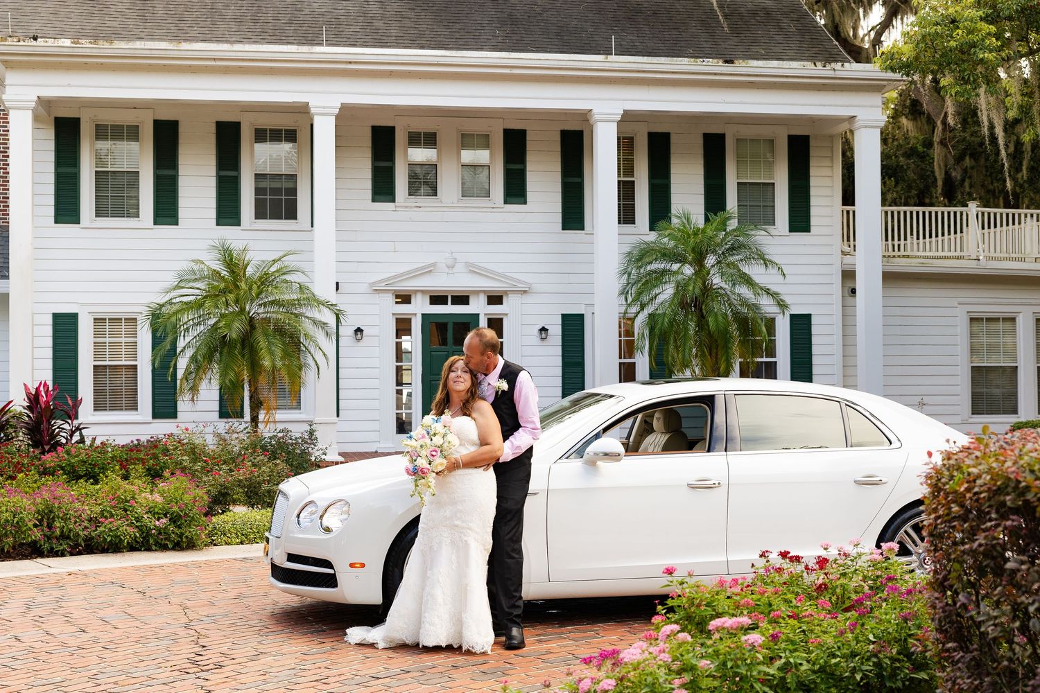 Bride and groom pose beside a white Bentley luxury car in front of elegant Southern mansion.
