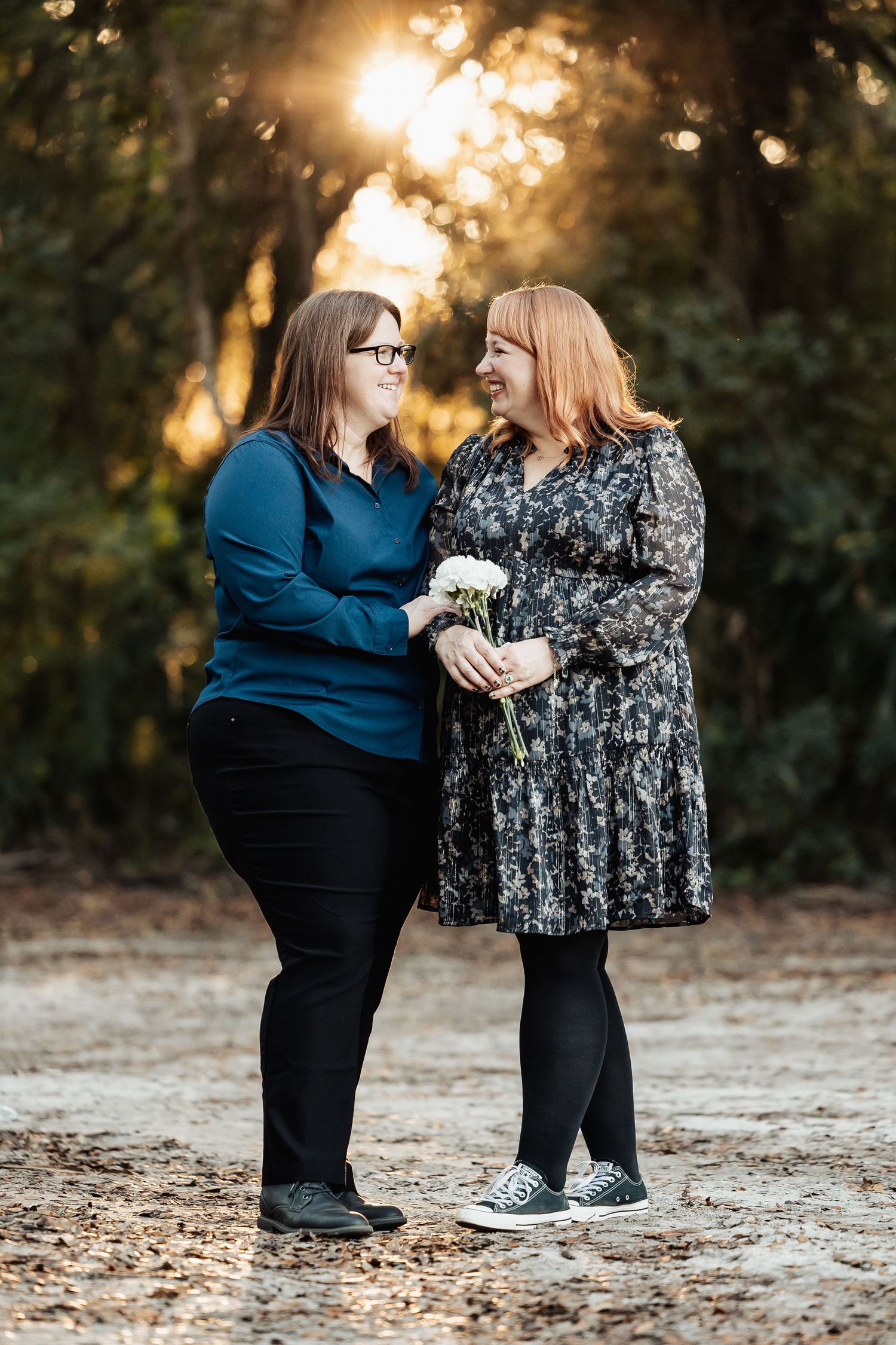 Two women smile at each other outdoors at golden hour, one holding white flowers during an engagement photo session.
