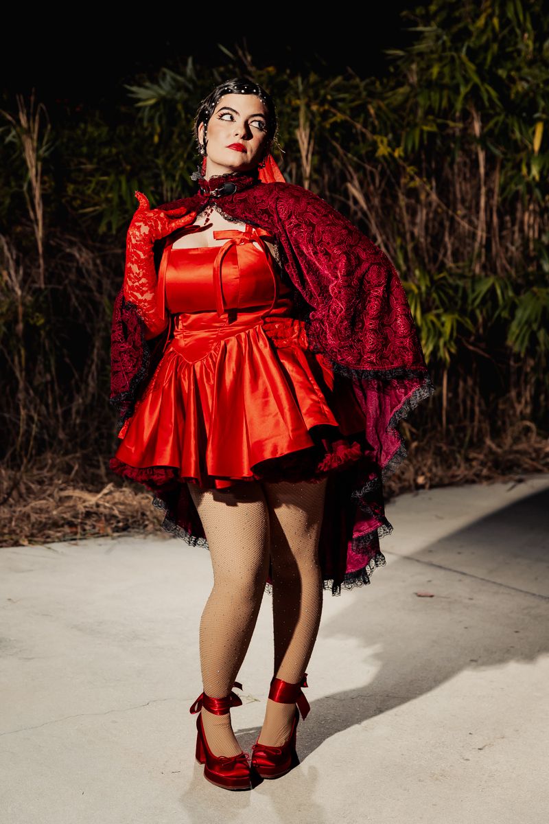 Woman in red satin mini dress with black lace cape posing outdoors at night wedding event.