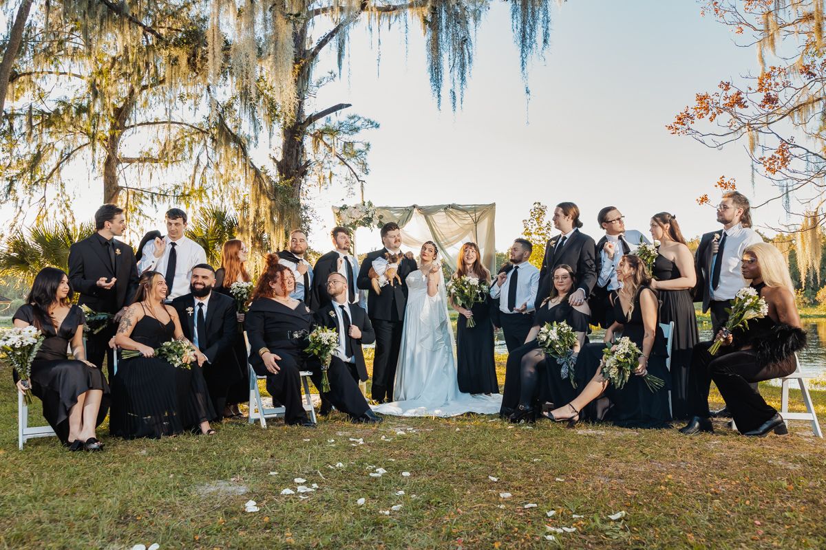 Wedding party in black attire posing with bride and groom under Spanish moss trees at sunset.