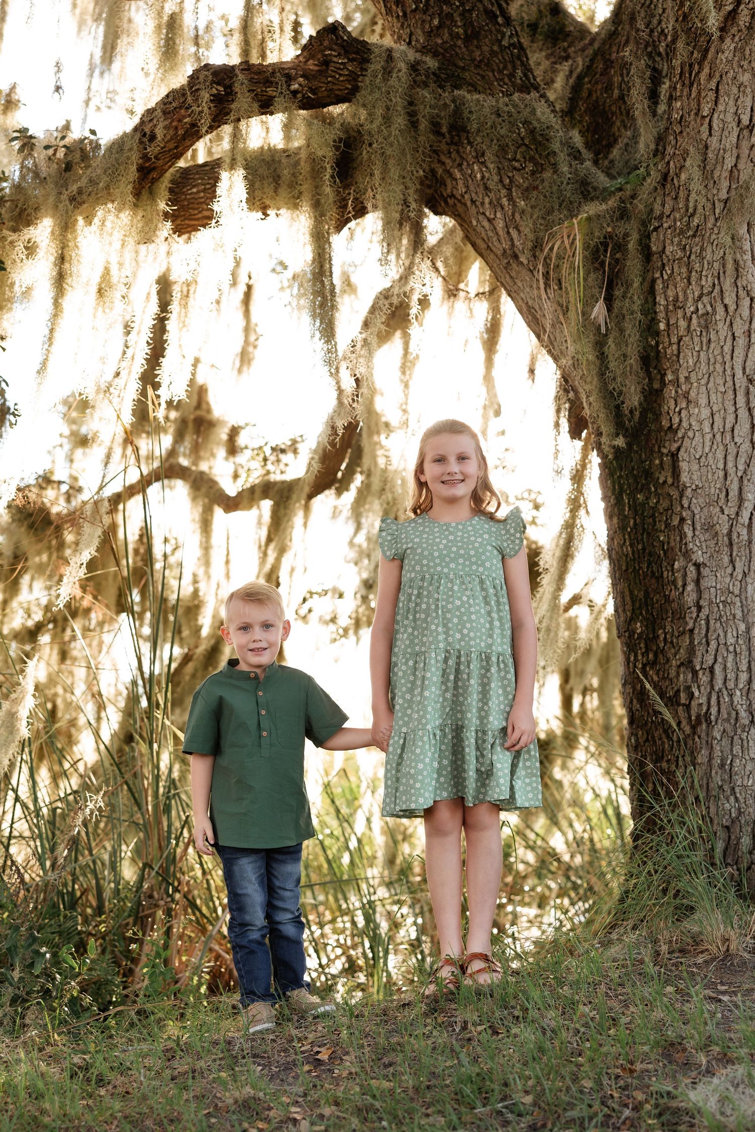 Two children stand together under a moss-draped tree in coordinating green outfits.