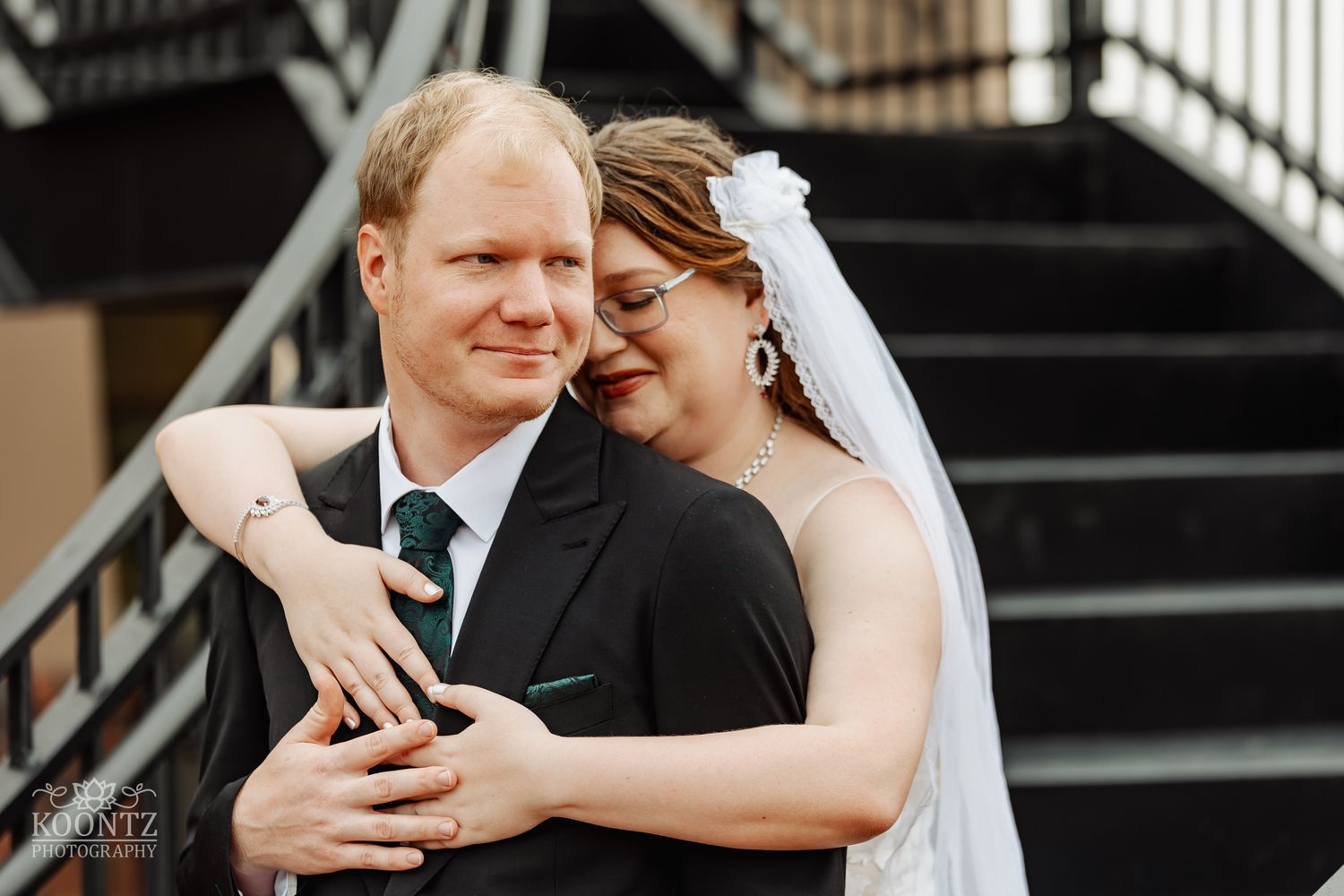 A tender embrace captured outdoors on a wedding day with a white bridal veil and dark suit.