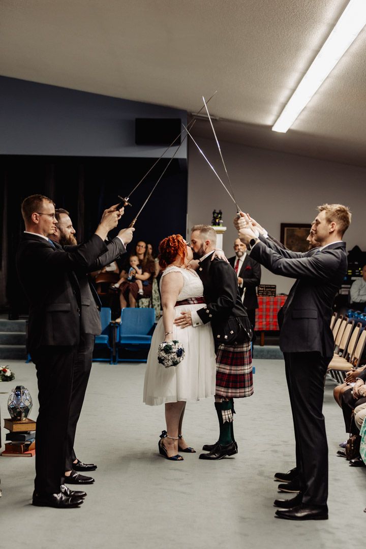 A wedding party forms a traditional sword arch during a Scottish-themed ceremony indoors.