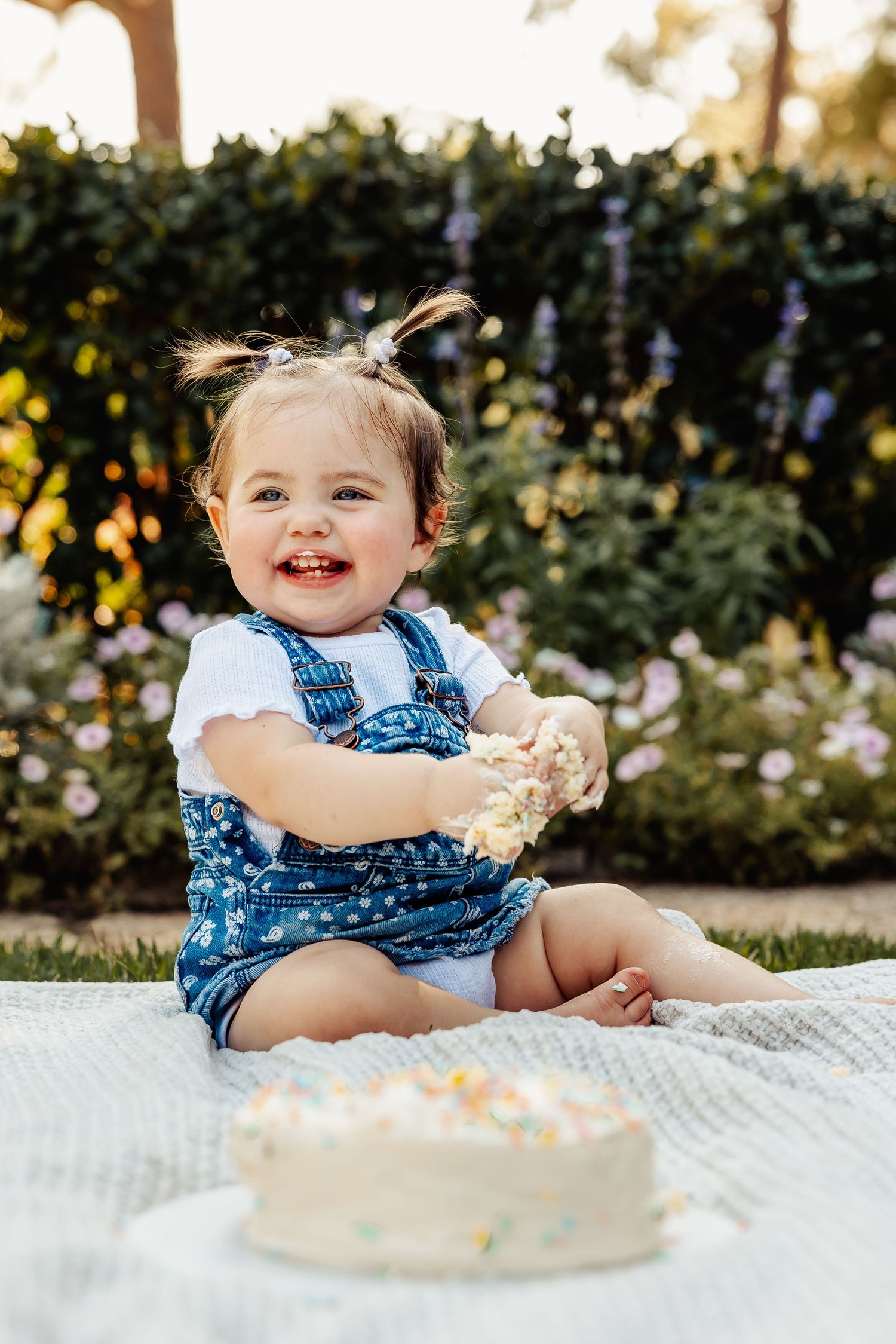Happy baby girl laughing and smashing her first birthday cake with frosting-covered hands outdoors.