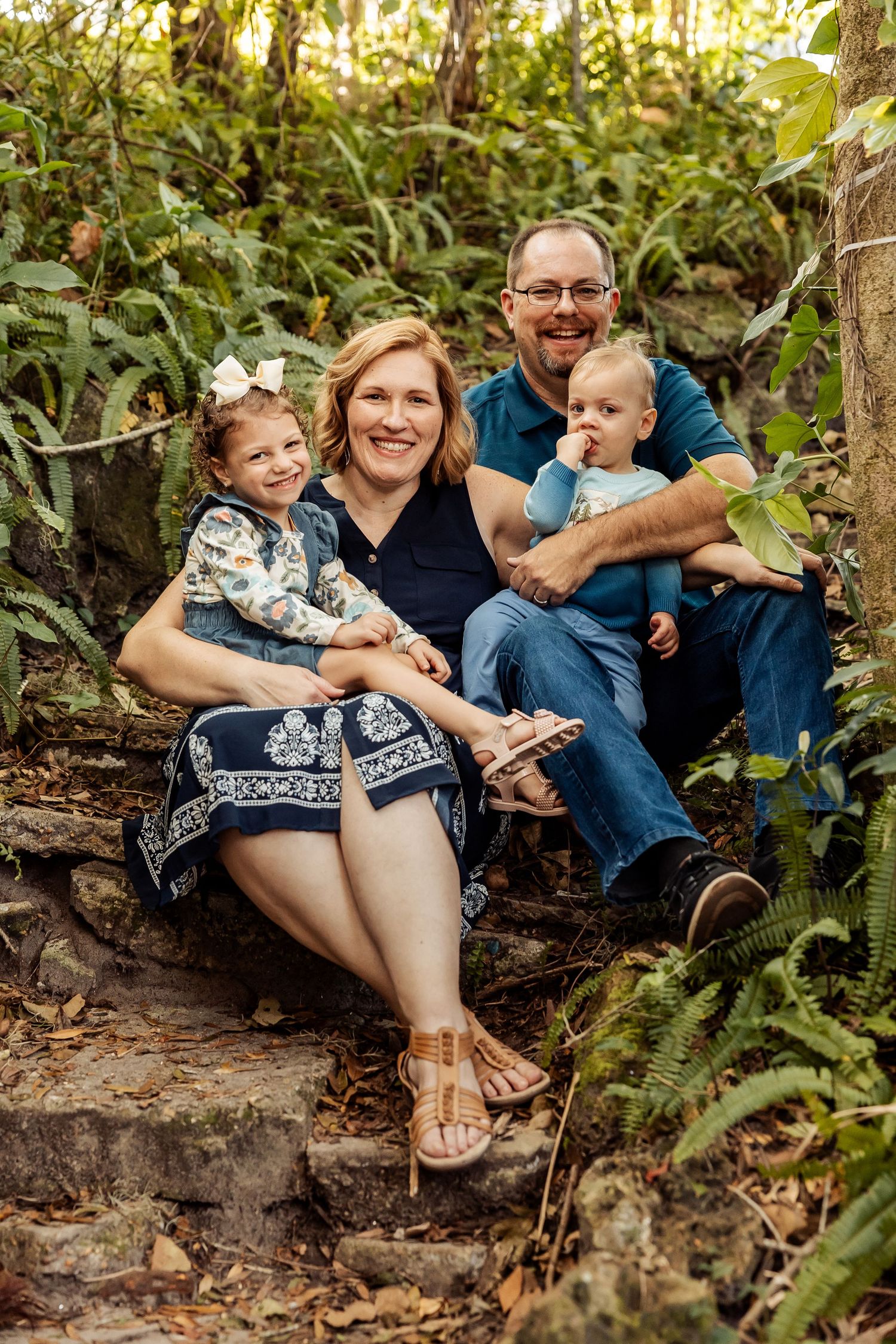 Happy family of four sitting on stone steps outdoors, parents holding two young children in a garden.