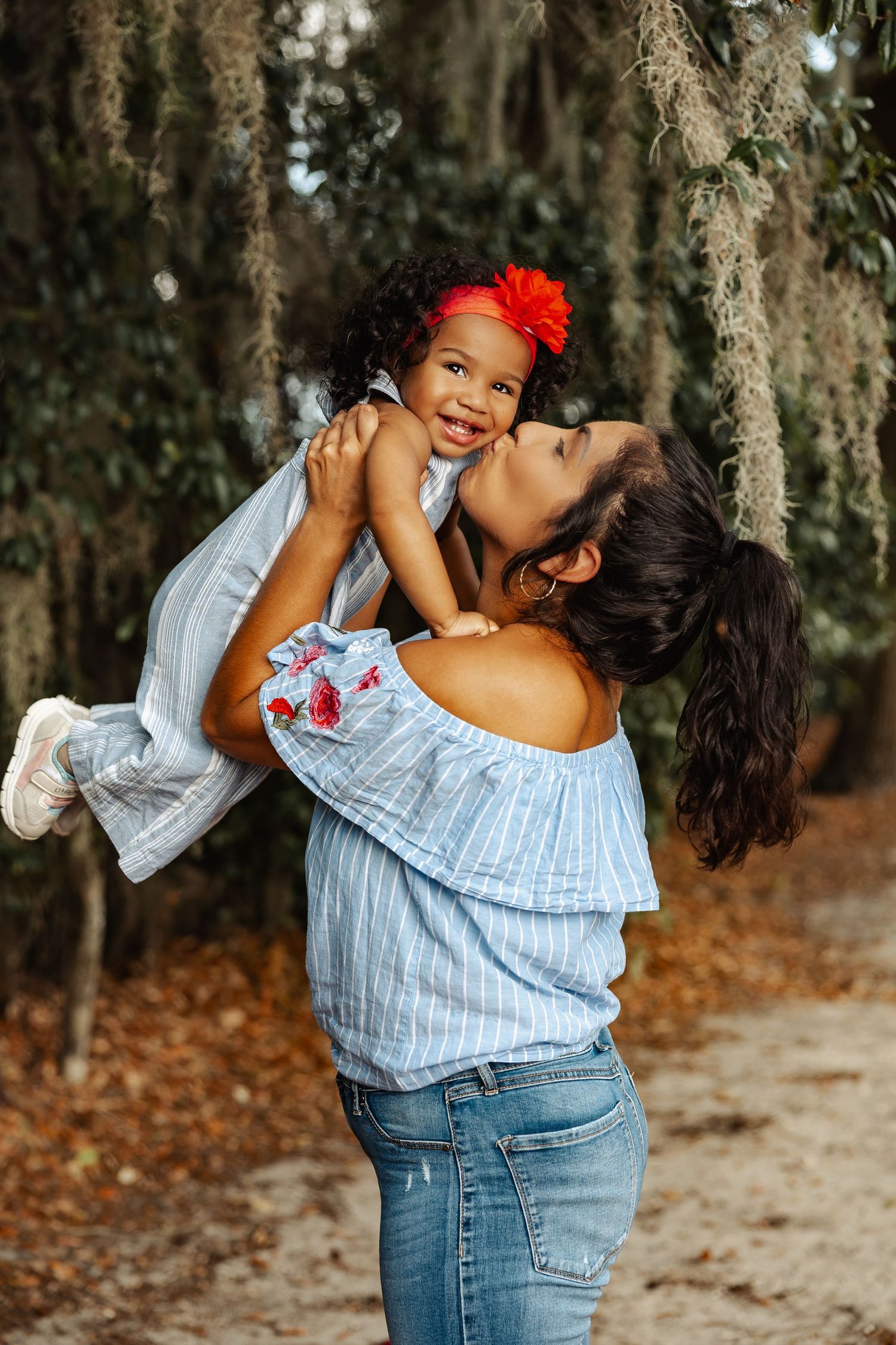 A mother kisses her smiling toddler daughter while lifting her among mossy trees.