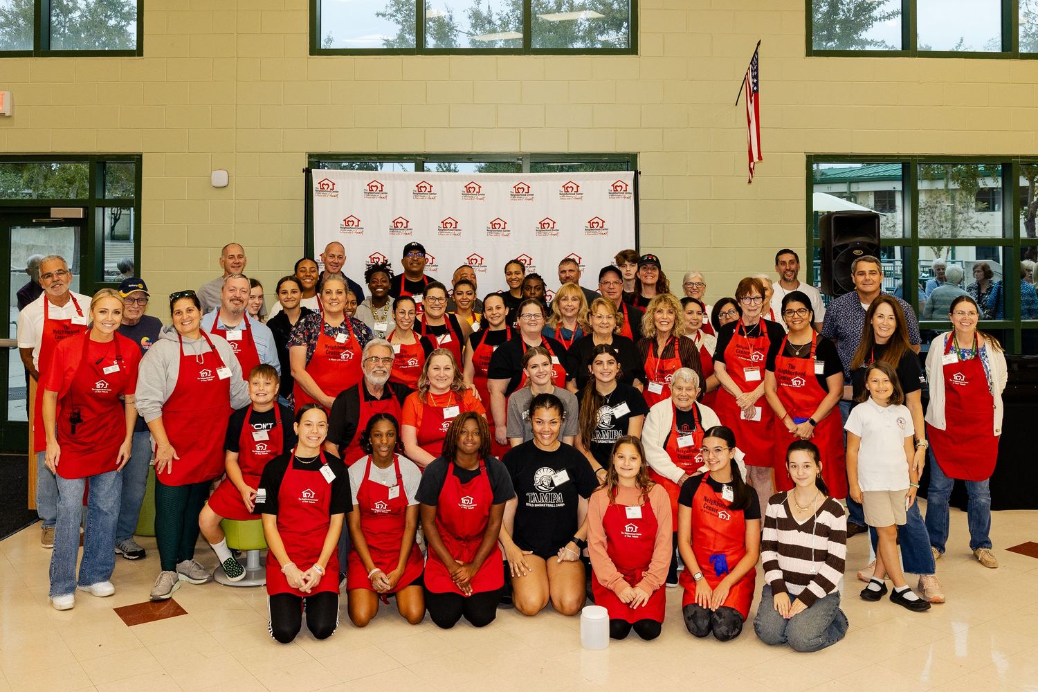Large group of volunteers in red aprons posing for a group photo in a school gymnasium.