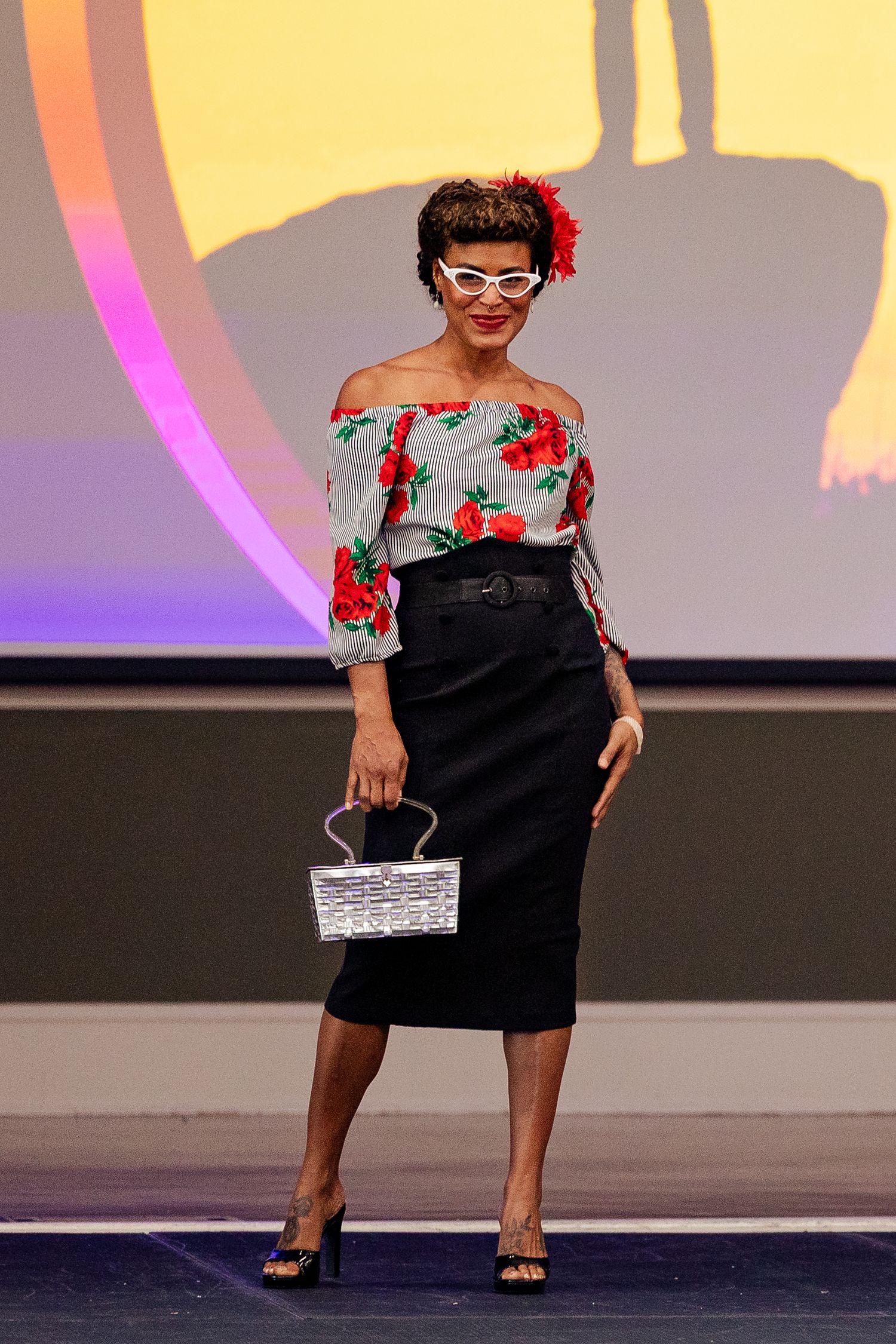 Woman poses in striped floral off-shoulder top and black skirt at fashion show.