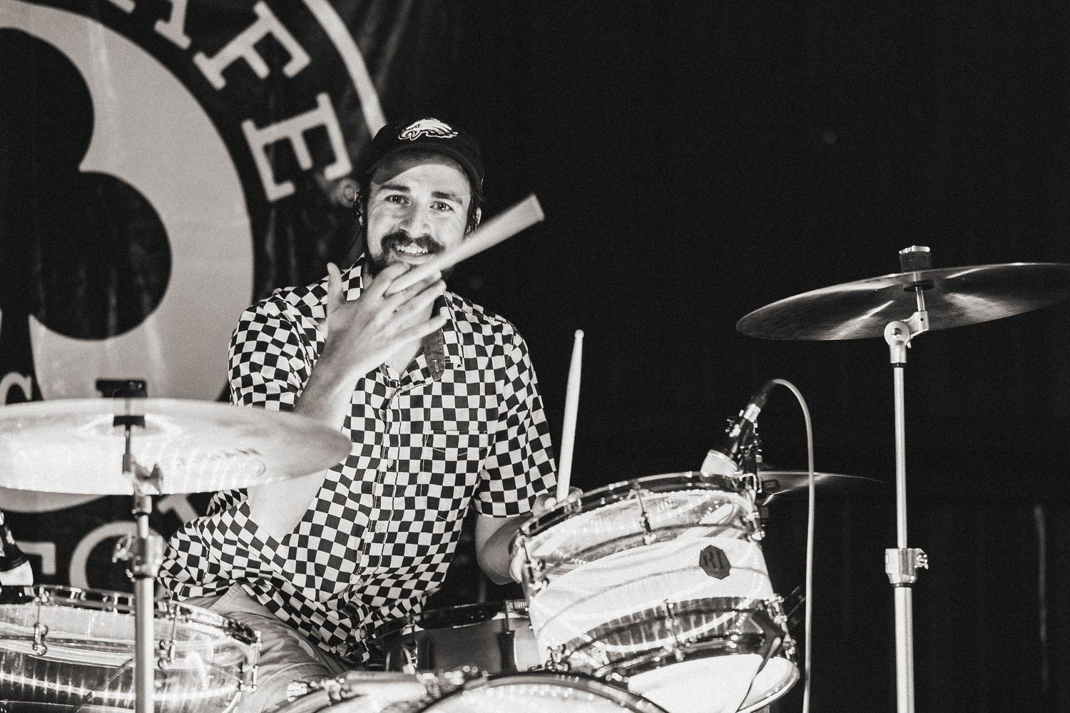 A bearded drummer in a checkered shirt plays drums in a vintage-style black and white concert photograph.