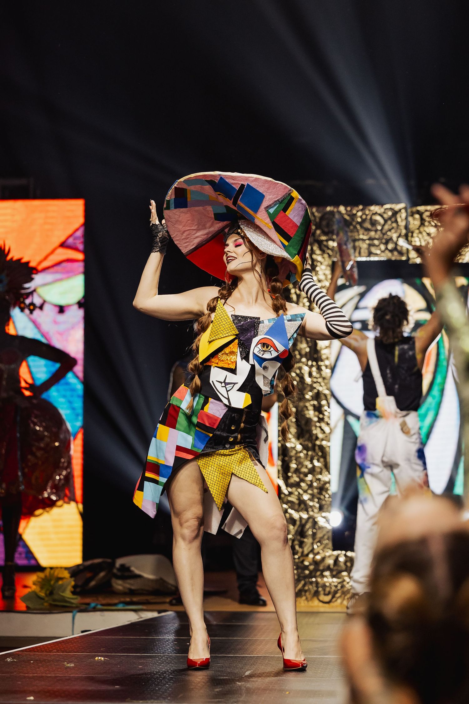 Model wearing colorful patchwork outfit with large hat on fashion runway stage.