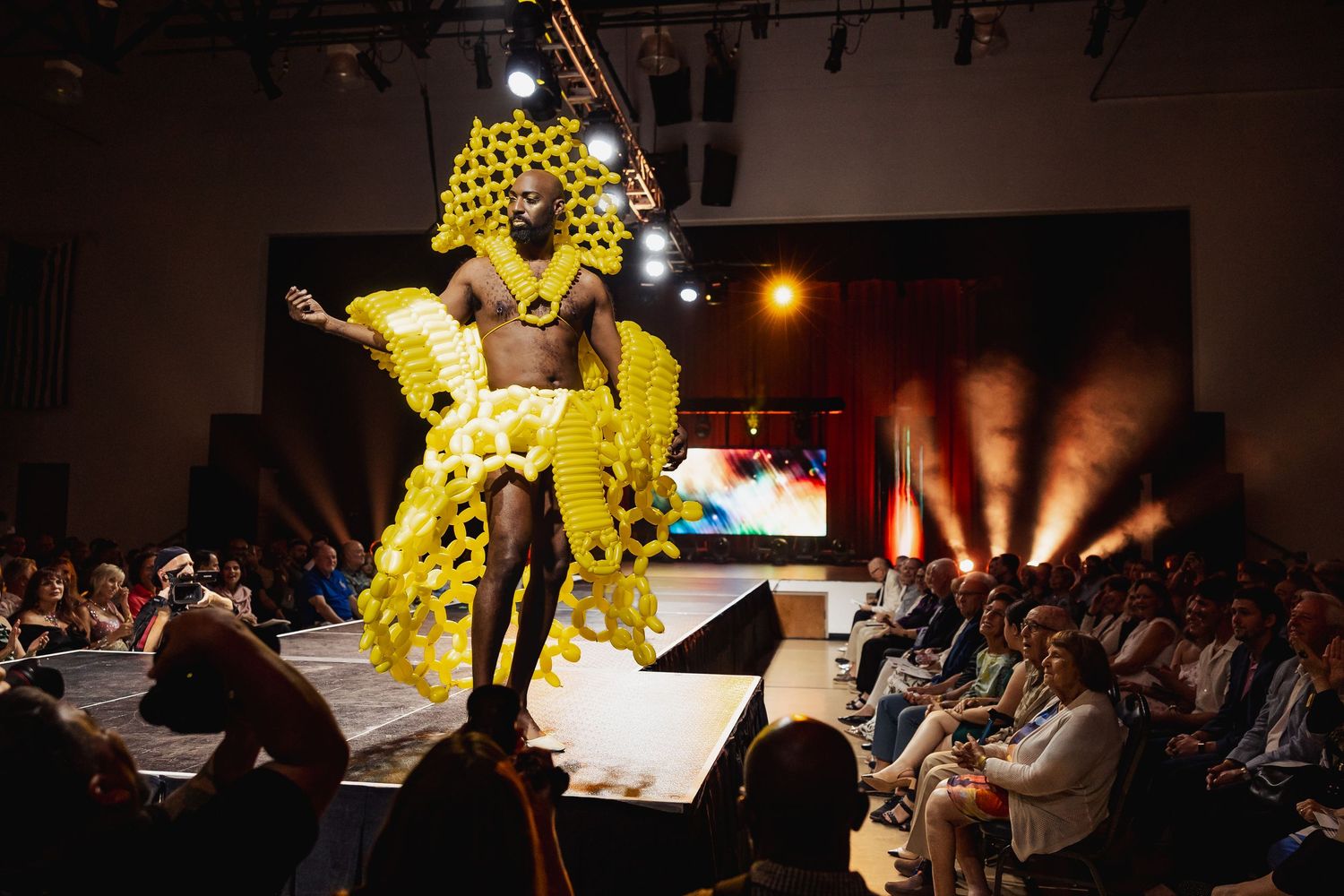 Model in yellow balloon gown strikes pose on runway before seated audience at balloon fashion show.