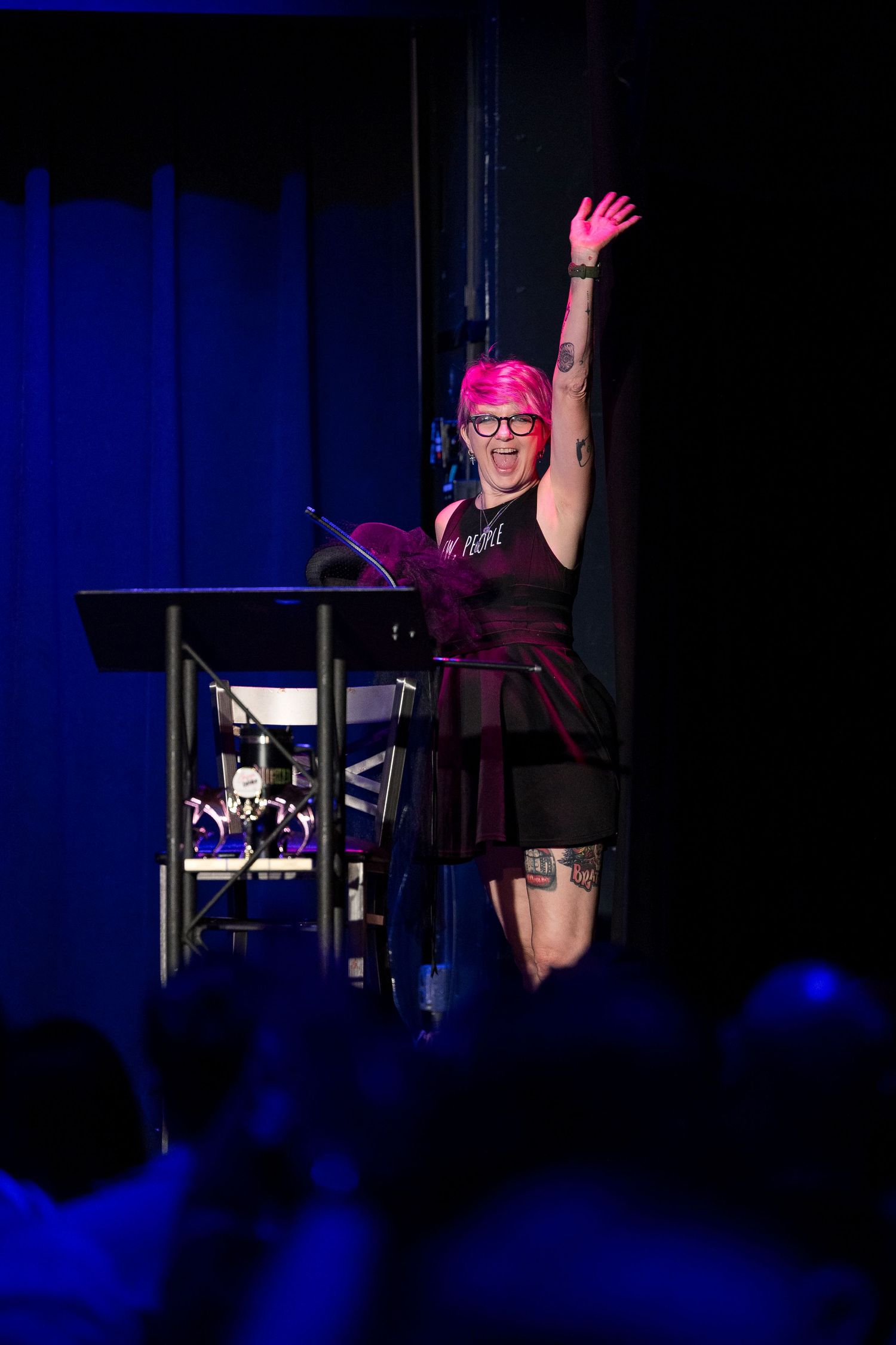 Pink-haired emcee with raised arm at podium under blue stage lighting at performance event.