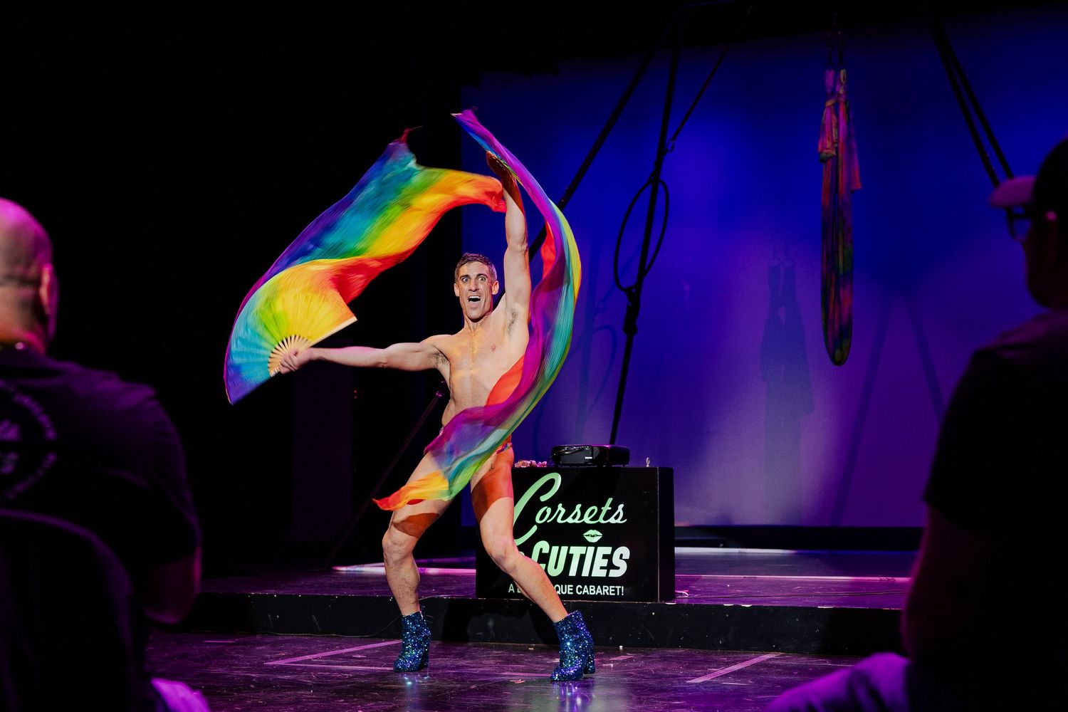 Shirtless male performer waving rainbow flag on stage at Corsets and Cuties burlesque cabaret show.