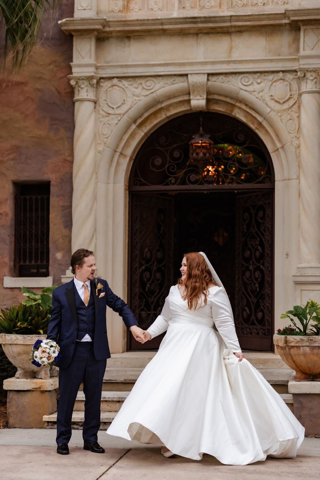 Bride in white gown and groom in navy suit hold hands, walking before ornate arched doorway at elegant wedding venue.