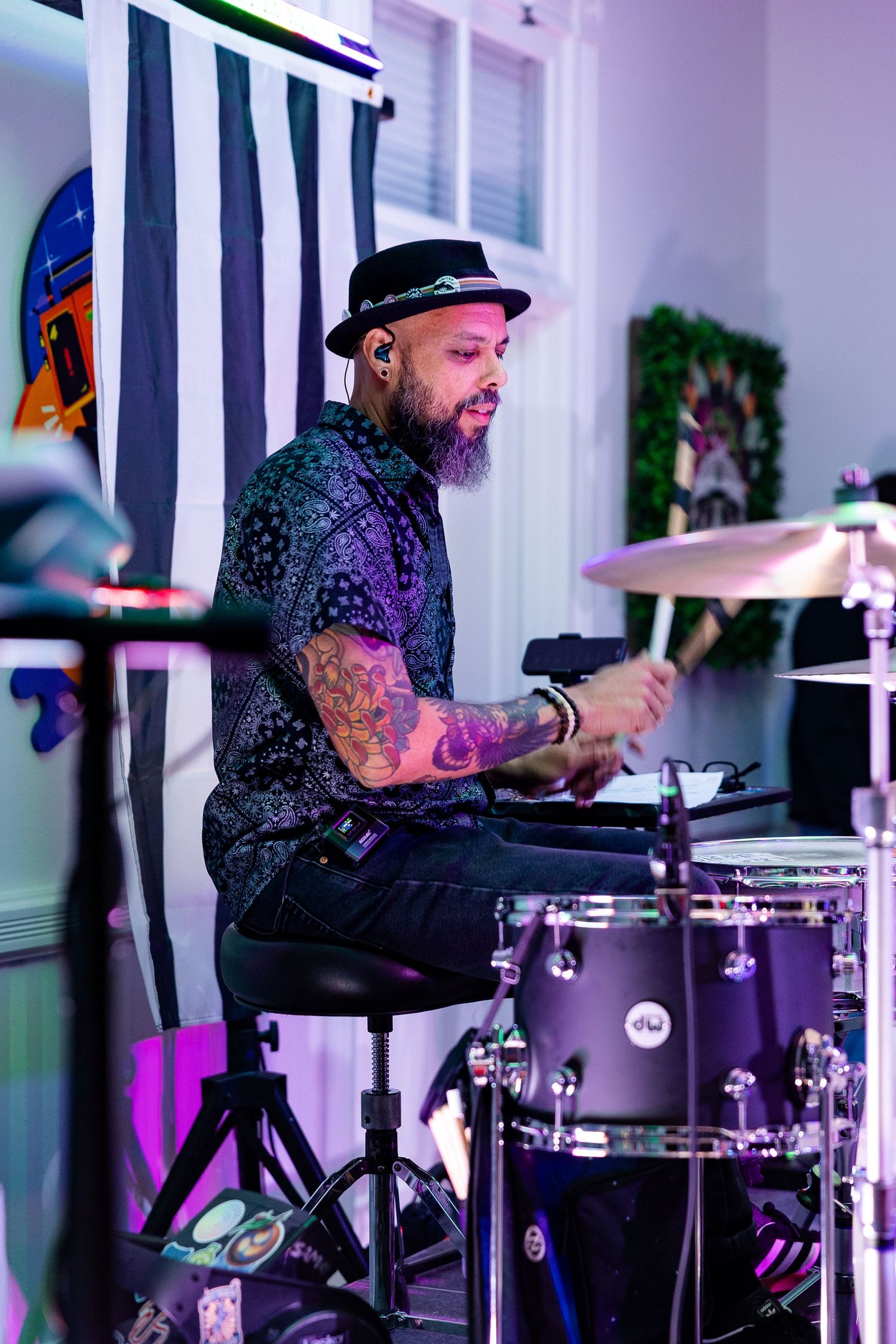Tattooed male drummer wearing a black hat plays drums on stage under purple lights.