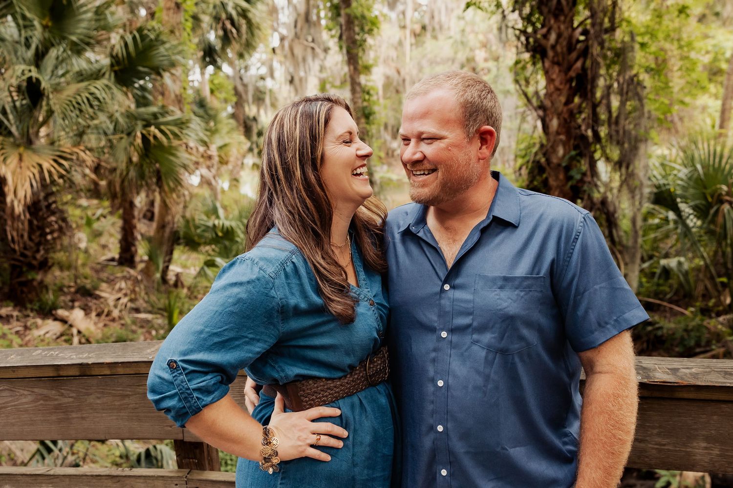 Smiling couple in blue outfits laughing together outdoors among lush green trees.