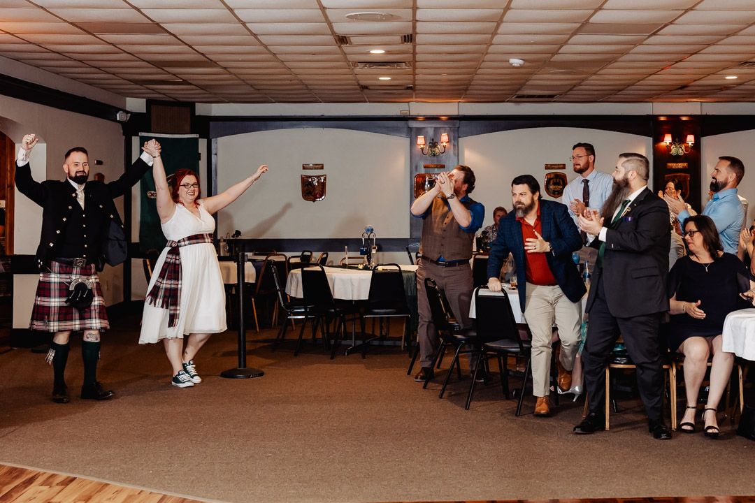 Bride and groom in kilt celebrating with raised hands at wedding reception with guests applauding.