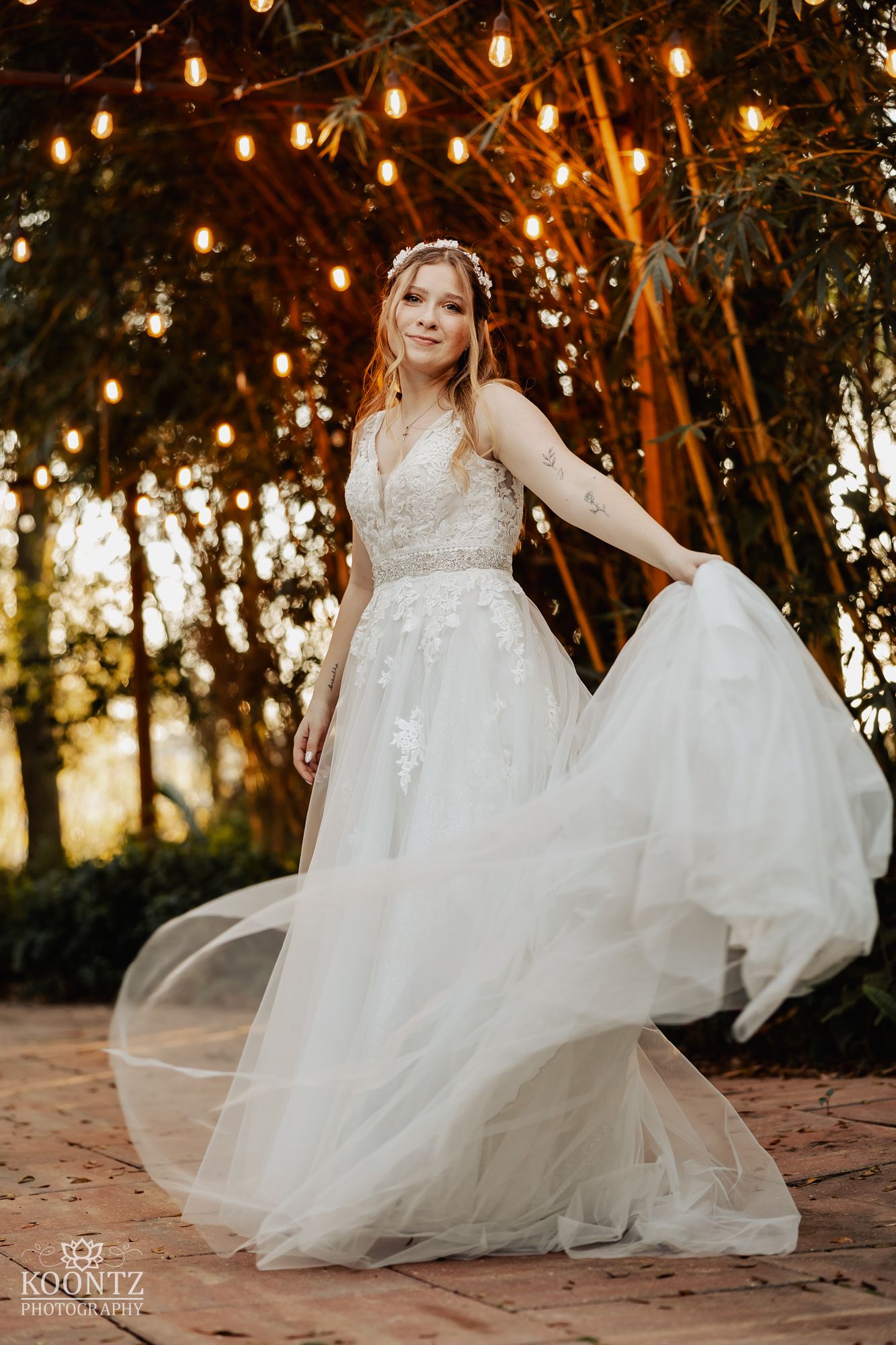 Bride in flowing lace tulle wedding gown twirls under warm string lights surrounded by bamboo trees at sunset.