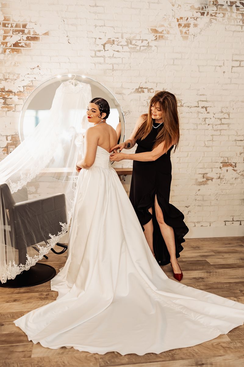 Assistant helping bride in elegant white gown with long train fasten her dress buttons.