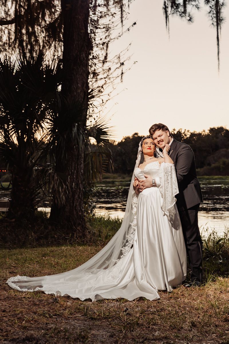 A couple shares a romantic moment under moss-draped trees at sunset by a lake, with the bride in a flowing white gown.