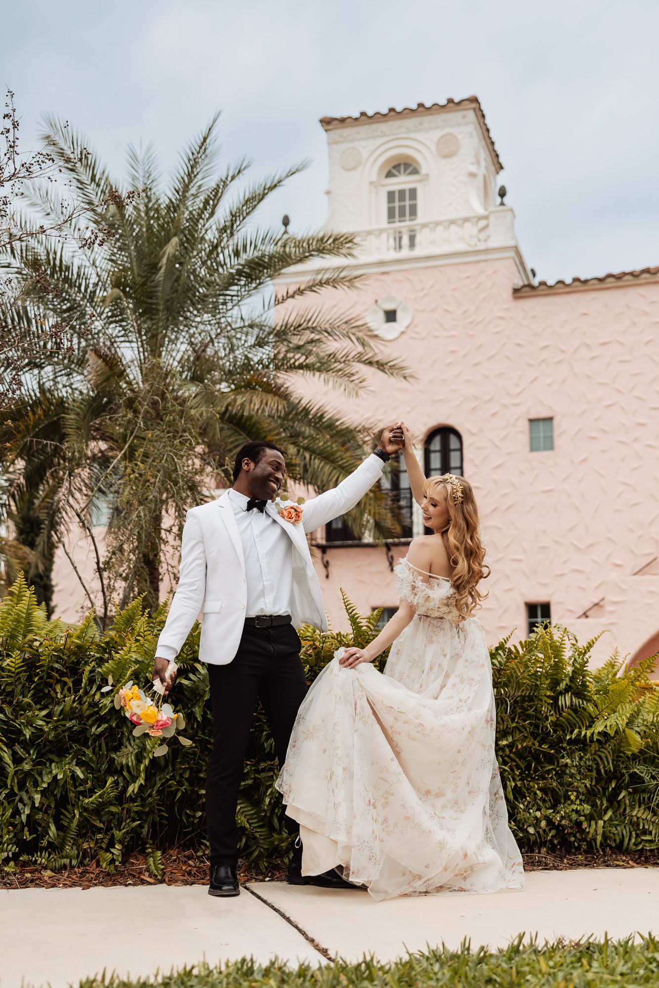 A couple in wedding attire dance together outside a Mediterranean-style building with a palm tree and tower.