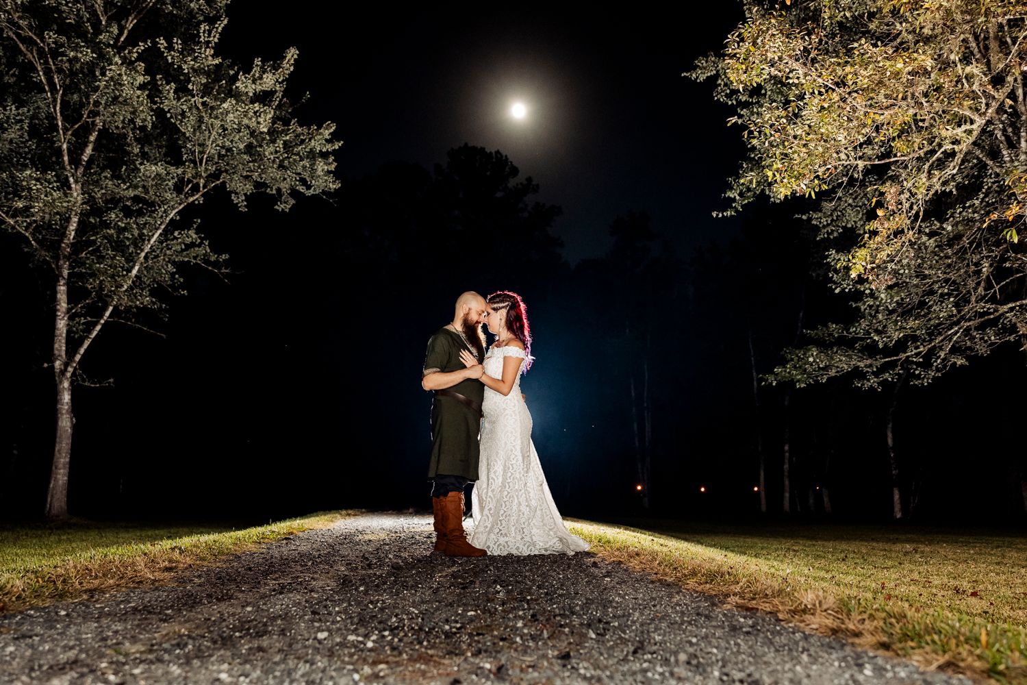 A romantic evening wedding portrait with a full moon and illuminated trees in the background.