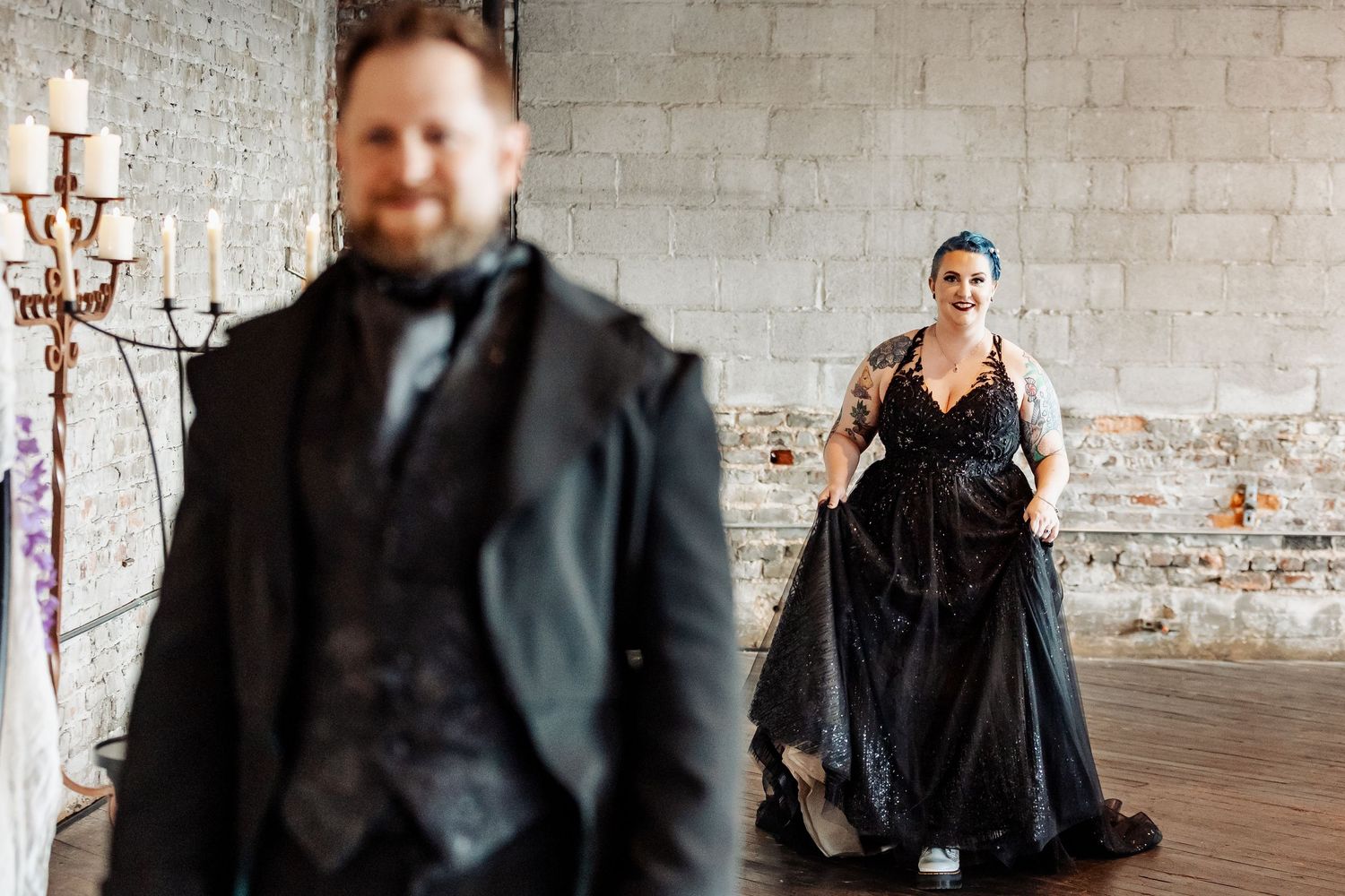 Tattooed bride in black ball gown smiling at groom in black suit in rustic brick venue.