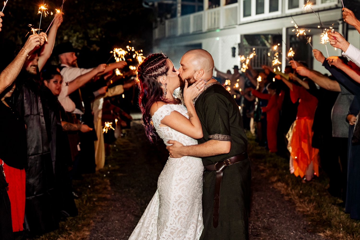 Bride and groom sharing passionate kiss during sparkler send-off exit surrounded by cheering wedding guests at night.