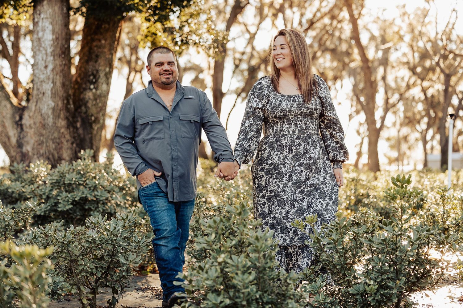 A happy couple walks hand in hand through a lush green park during their engagement session.