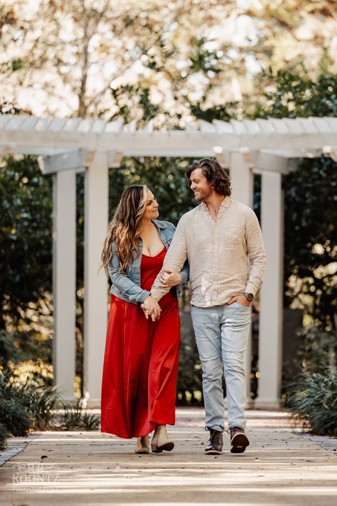 A couple walking hand in hand through a white columned gazebo in a garden setting.