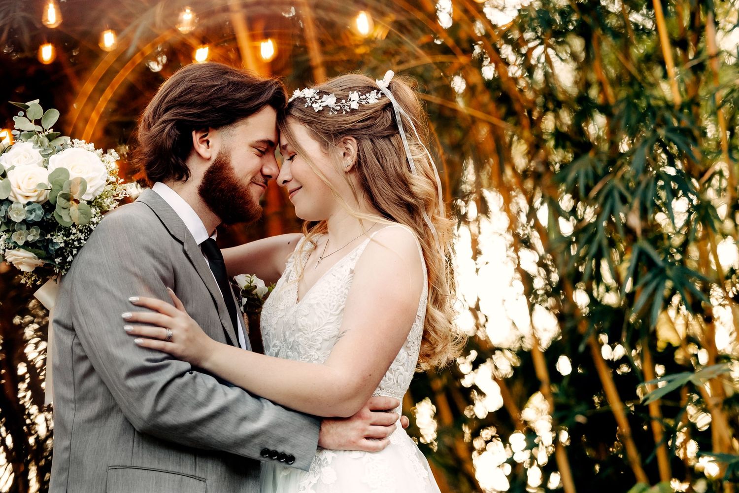 A romantic outdoor wedding portrait with bokeh lights and greenery in the background.