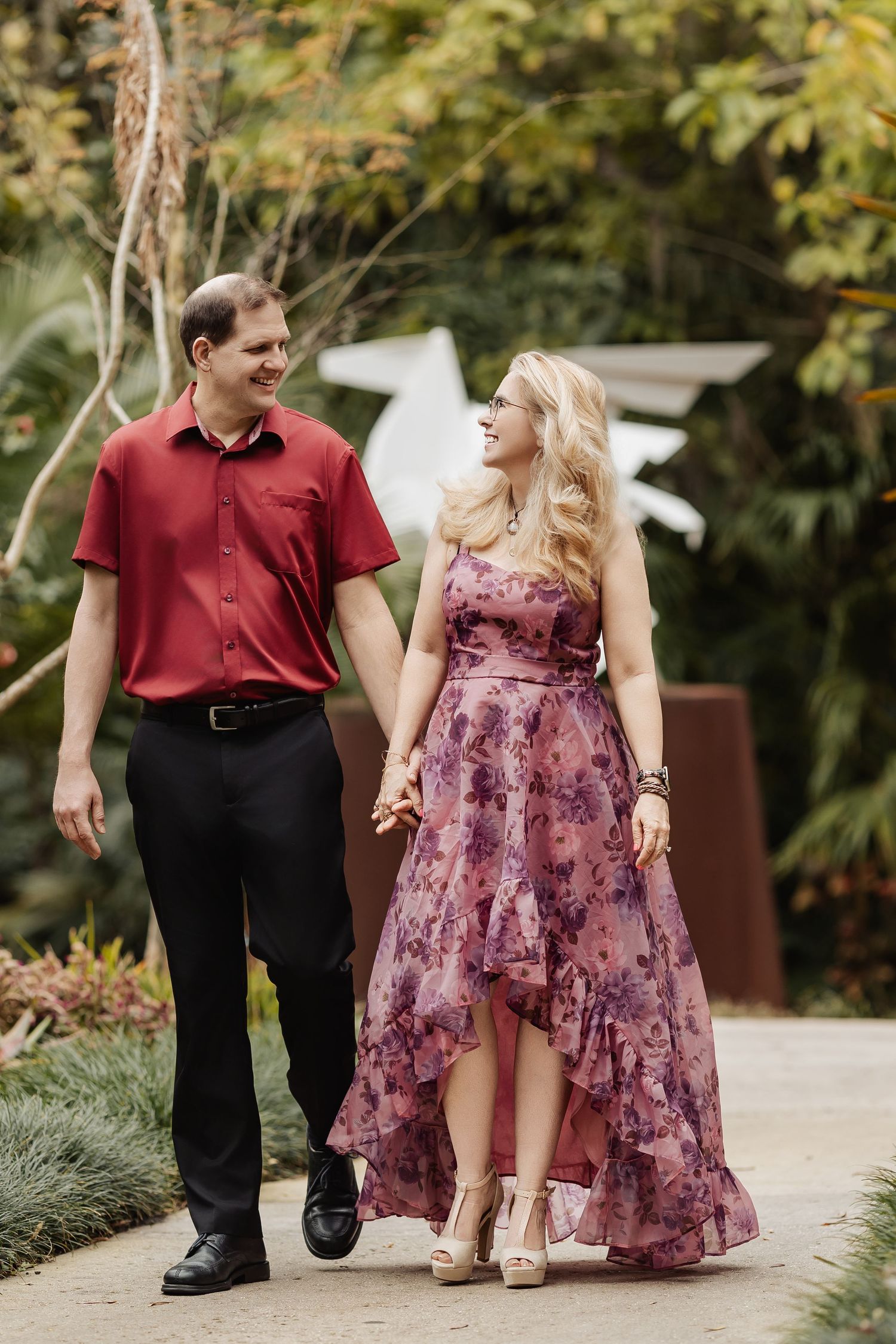 Happy couple walking hand in hand through lush garden in coordinating red and floral outfits.