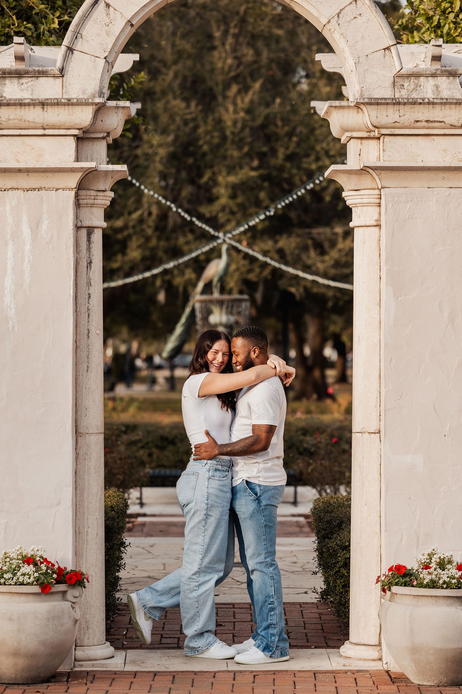 Couple sharing a warm embrace framed by ornate white stone archway with garden fountain backdrop.