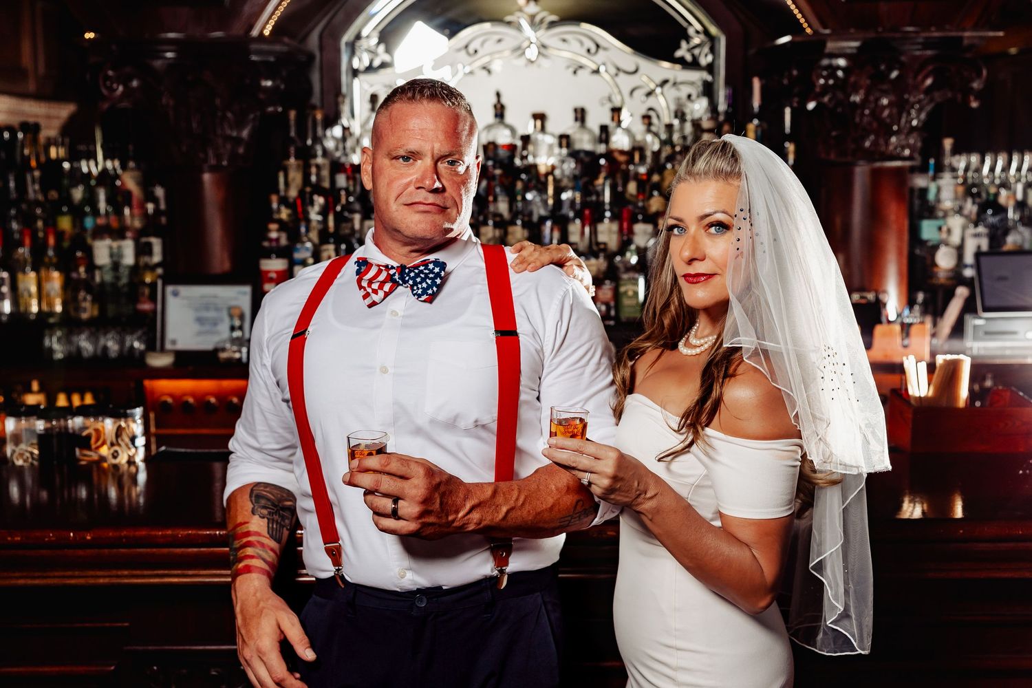 Bride and groom posing seriously at upscale bar, groom in red suspenders and patriotic bow tie.