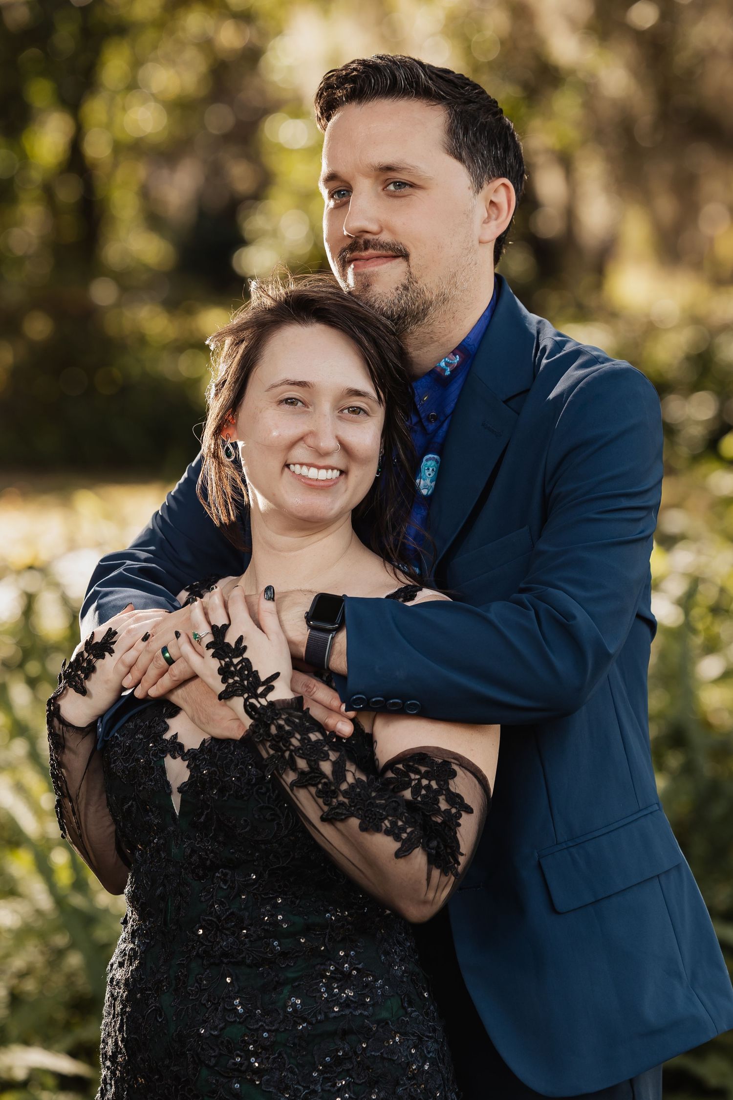 Smiling bride in black lace dress embraced by groom in blue suit in a sunlit outdoor garden setting.