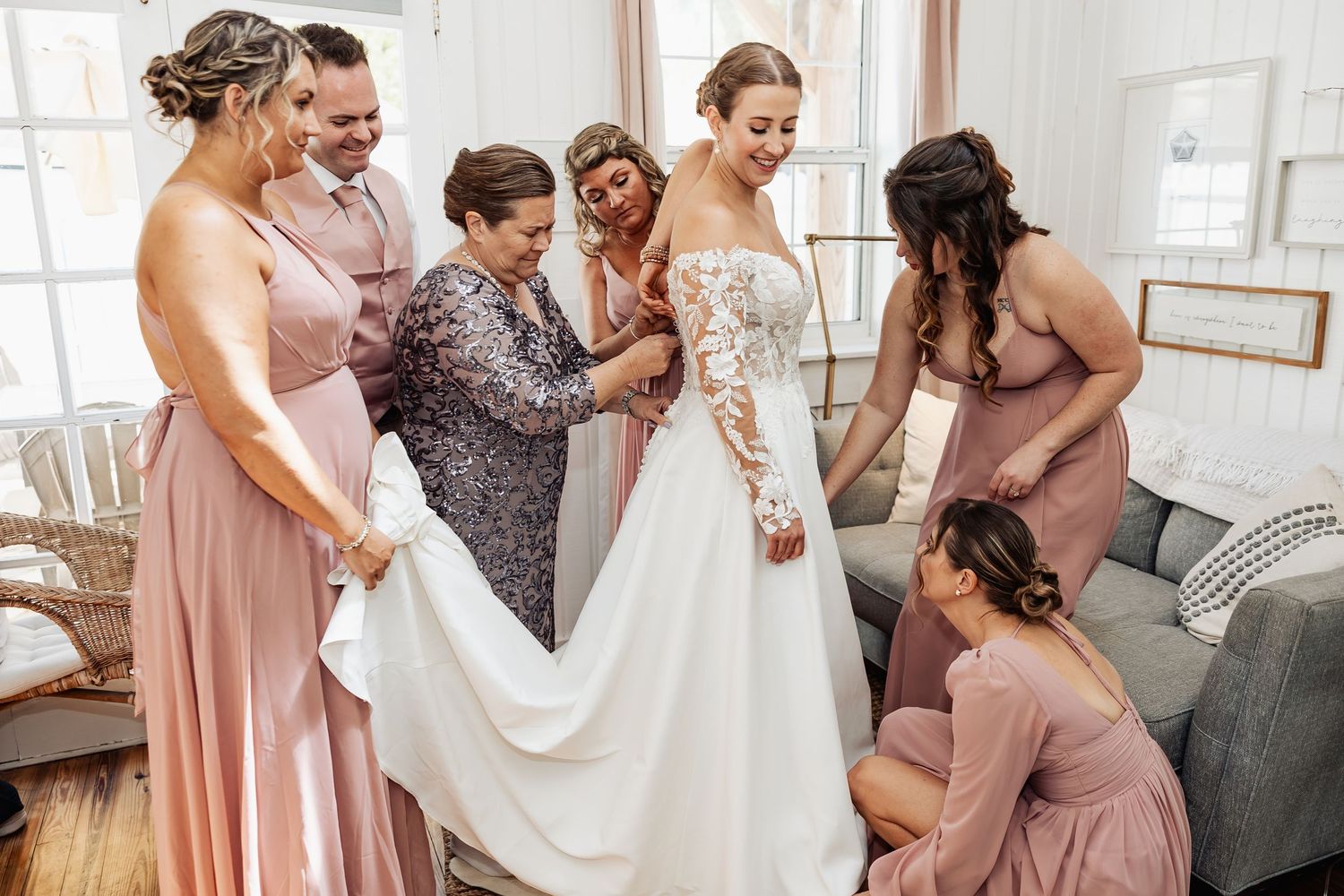 Bride in lace wedding gown surrounded by bridesmaids in dusty rose dresses being helped to dress.