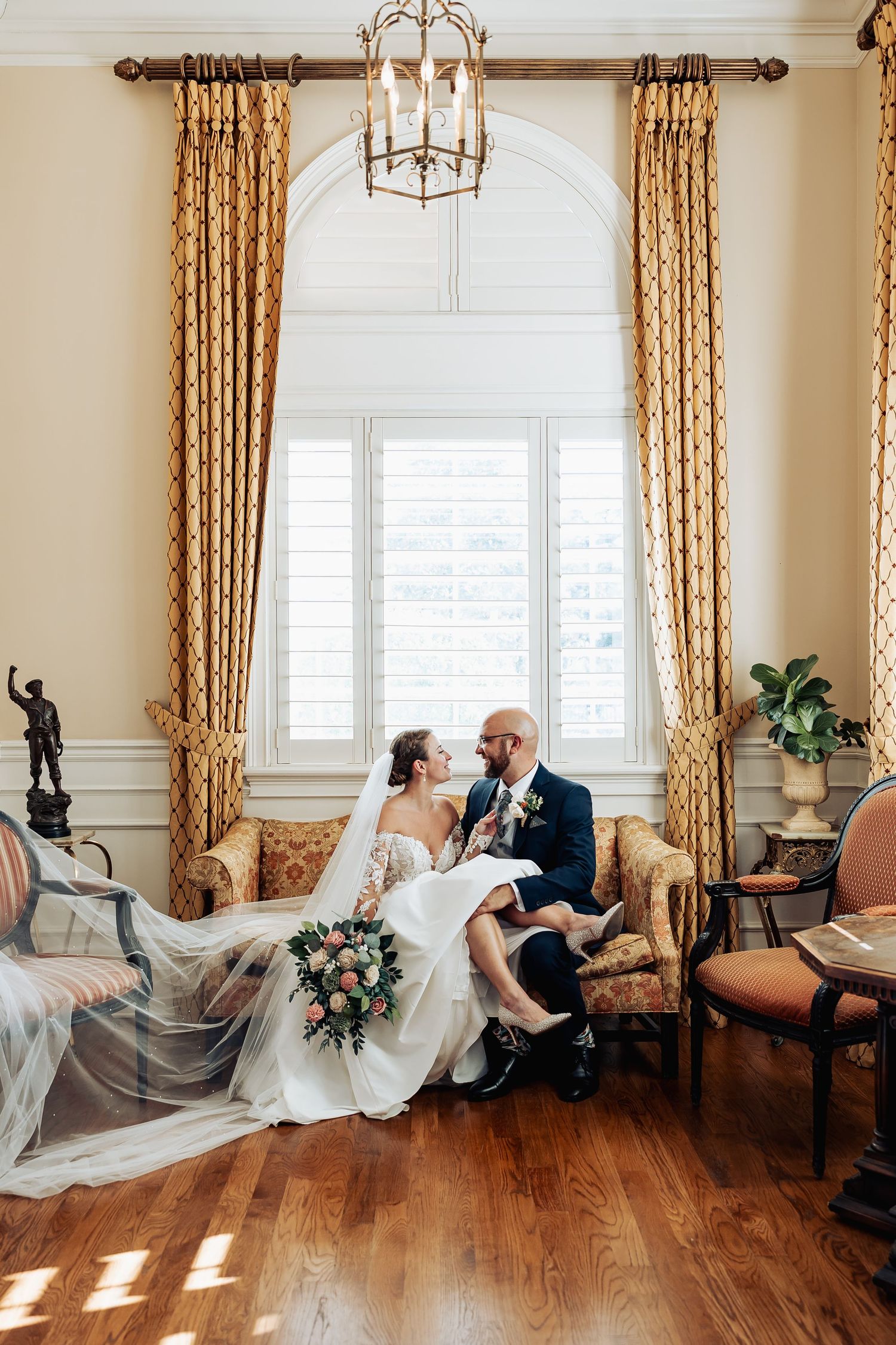 Bride and groom sitting together on elegant sofa with flowing veil in decorated room.