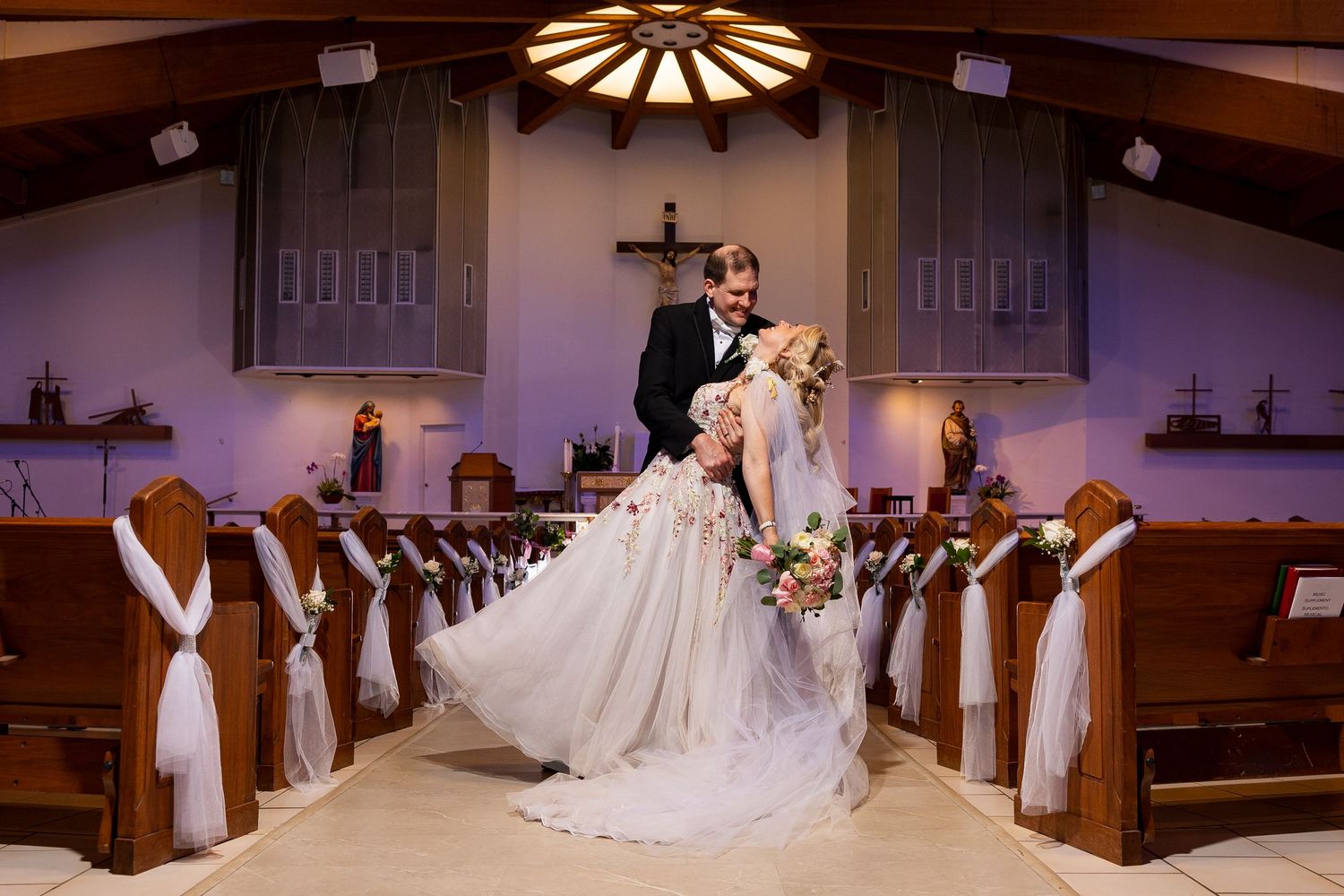 Groom lifting bride in ornate floral wedding dress in elegant church aisle with wooden pews.